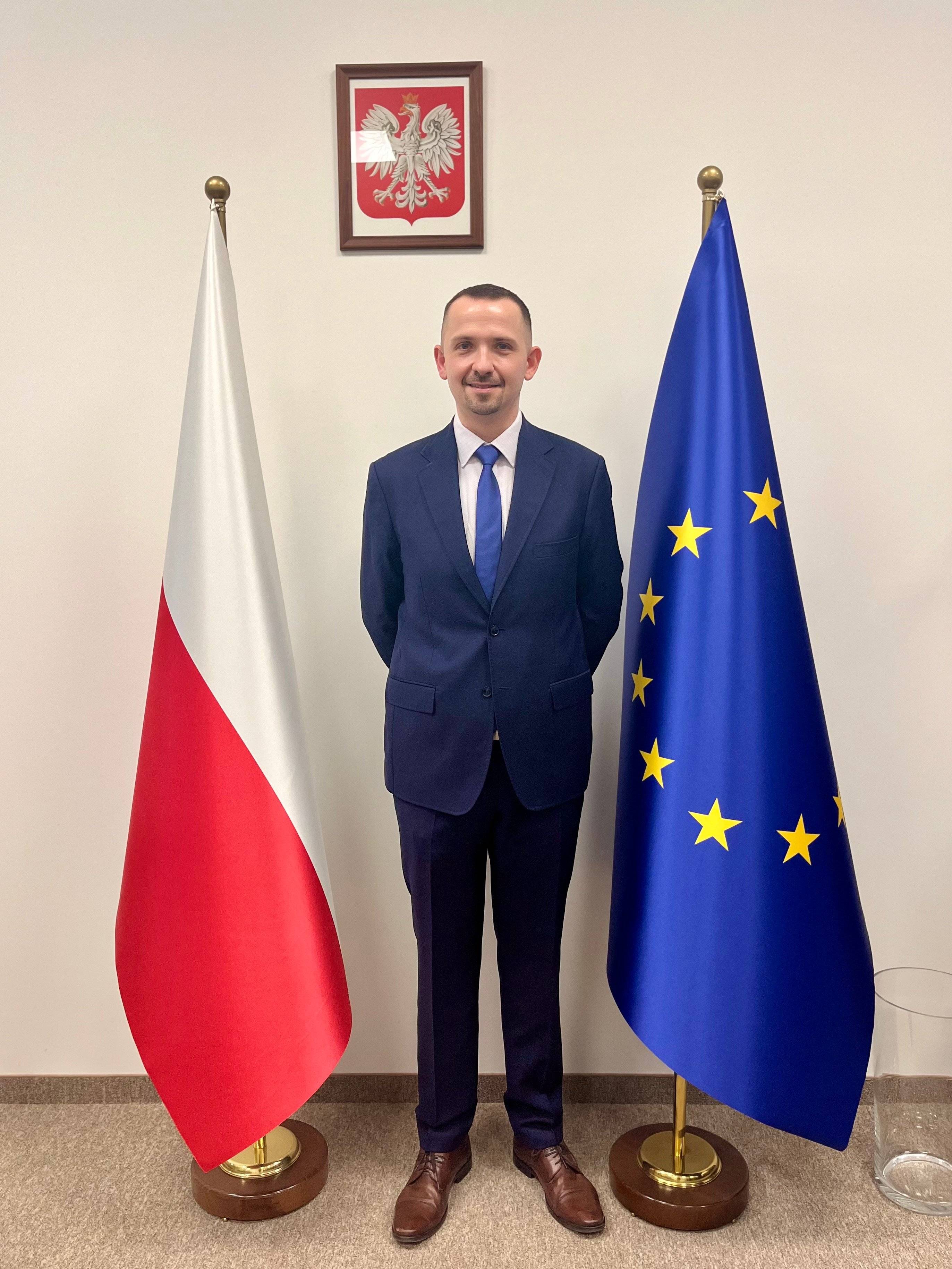 Młody męzczyzna w garniturze stoi między polską i unijna flagą, nad nim godło państwowe