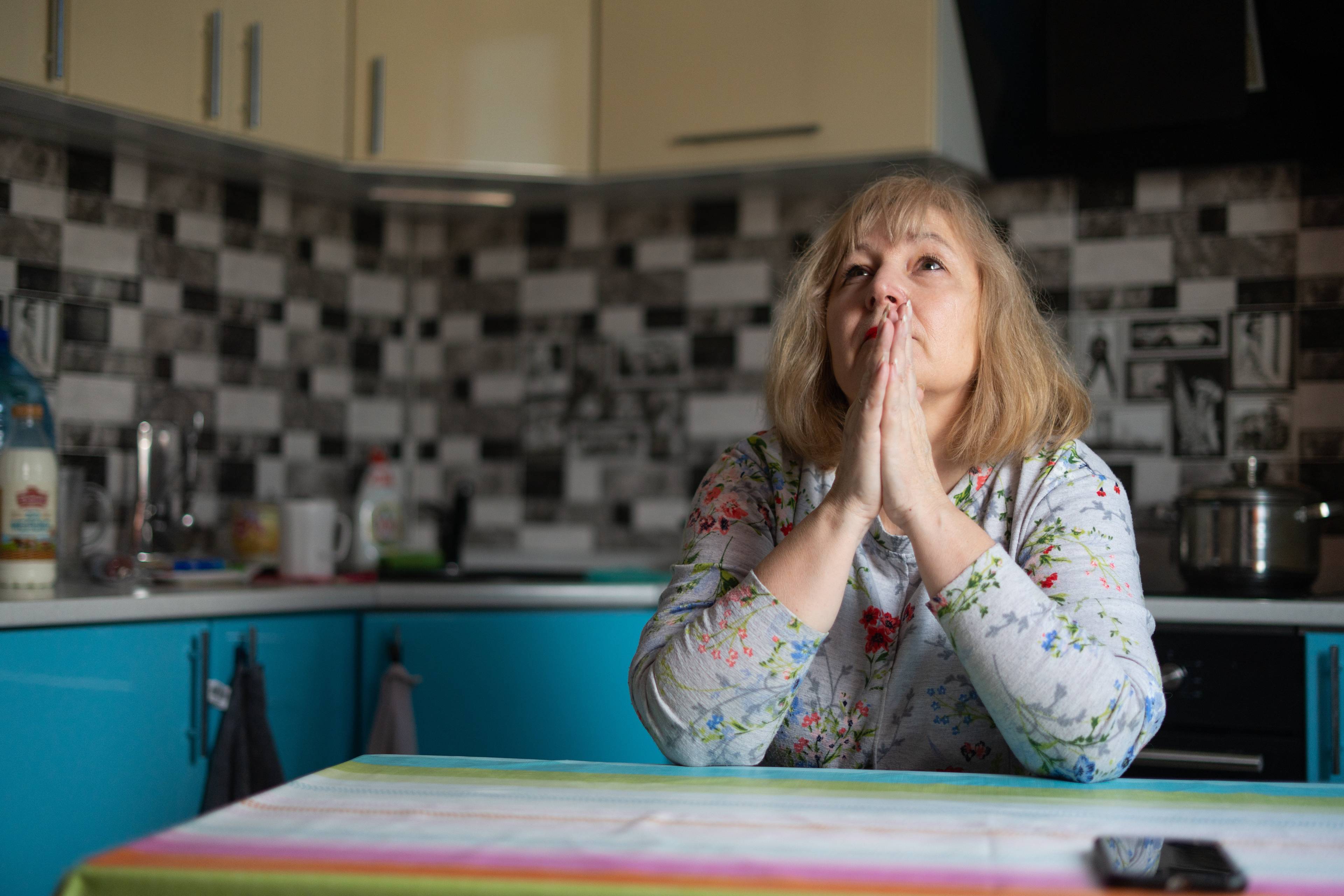 kobieta w średnim wieku z blond włosami do ramion siedzi w kuchni składając ręce jak do modlitwy