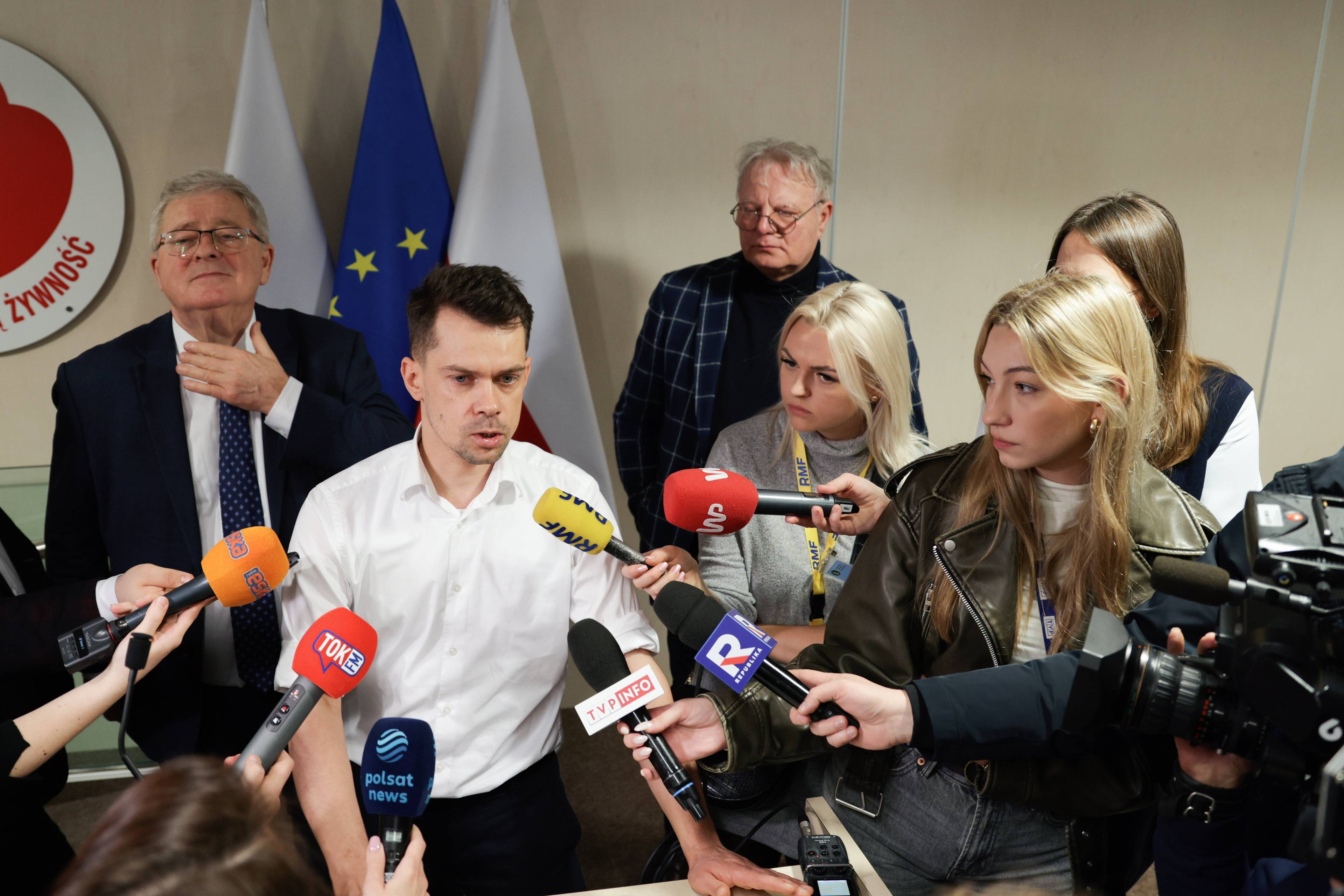 Michał Kołodziejczak w białej koszuli przed mikrofonami mediów podczas konferencji prasowej