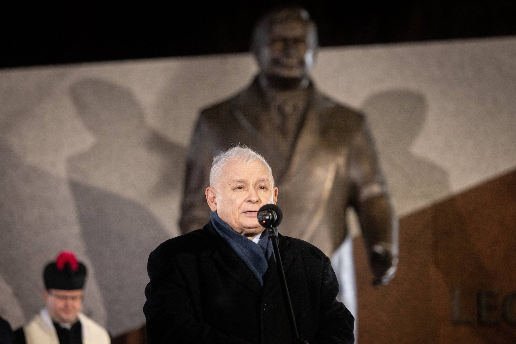 Jarosław Kaczyński przemawia na tle pomnika Lecha Kaczyńskiego. Ubrany w czarny płaszcz i granatowy szalik.