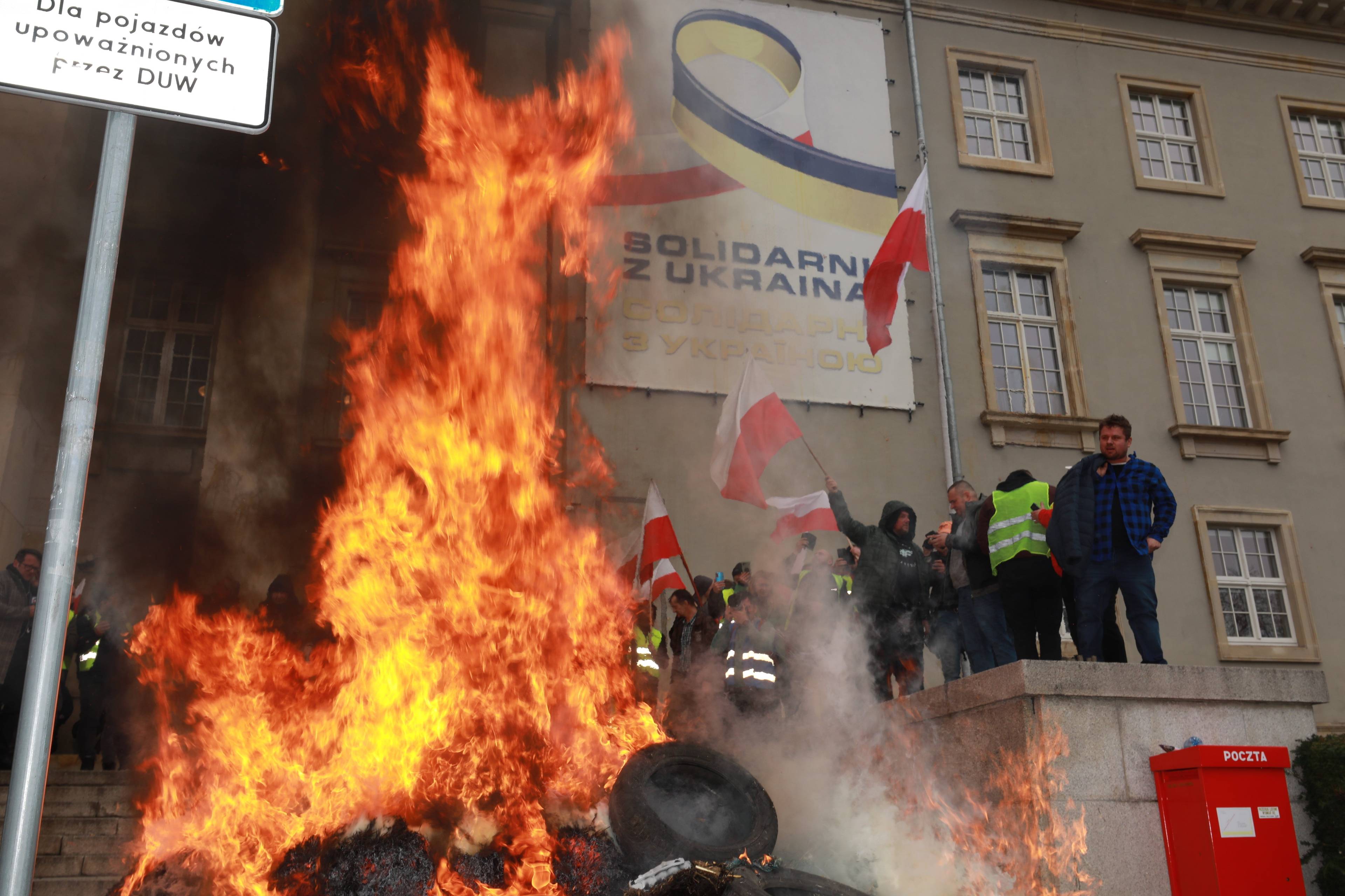 Na pierwszym planie płomienie. Na drugim: grupa osób z biało-czerwonymi flagami. W tle, na ścianie budynku, napis „Solidarni z Ukrainą”