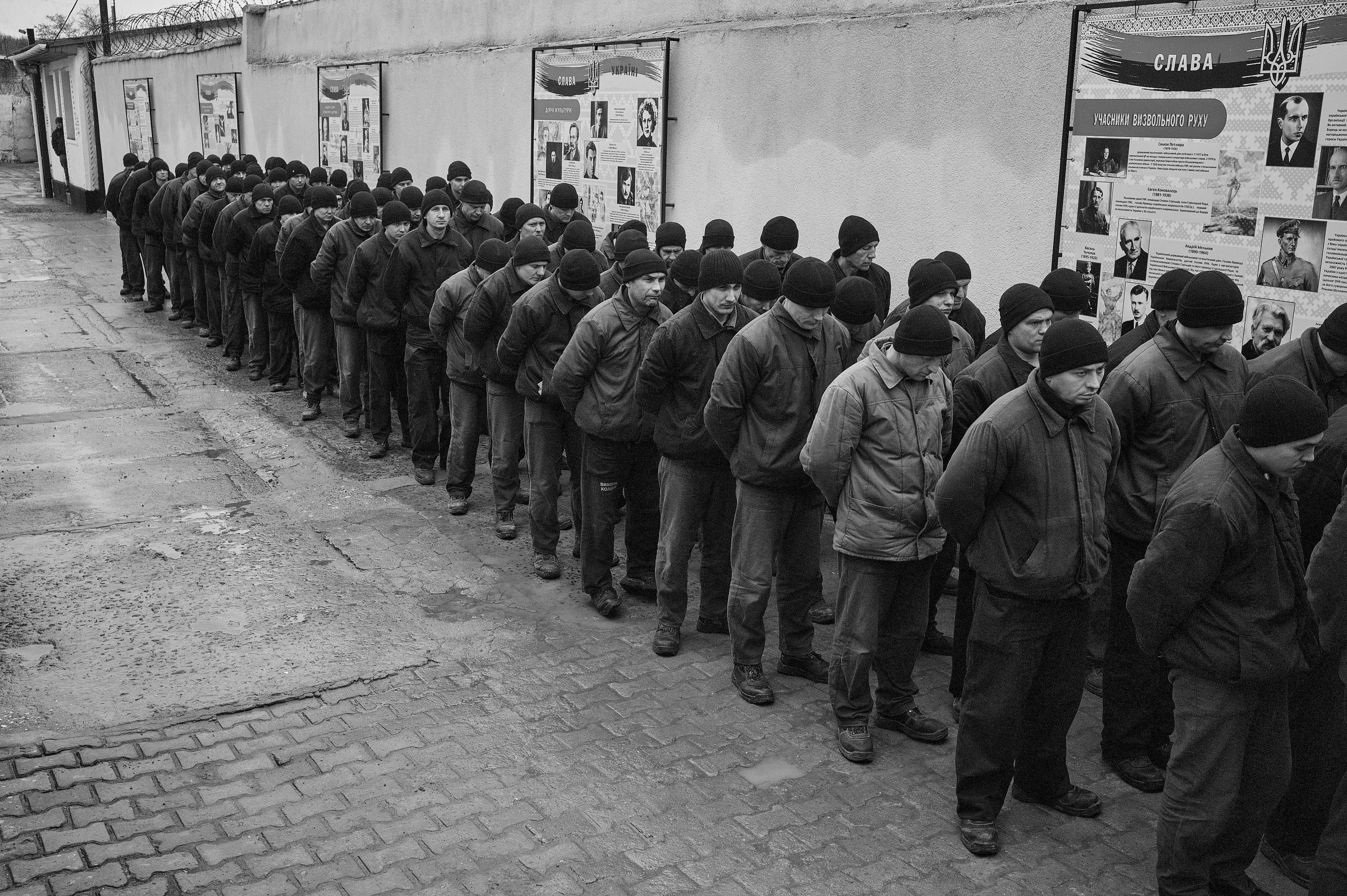 zdjęcie czarno-białe. Mężczyźni w ciemnych kurtkach i czarnych czapkach stoją trójkami pod ścianą, na której wiszą materiały o historii Ukrainy