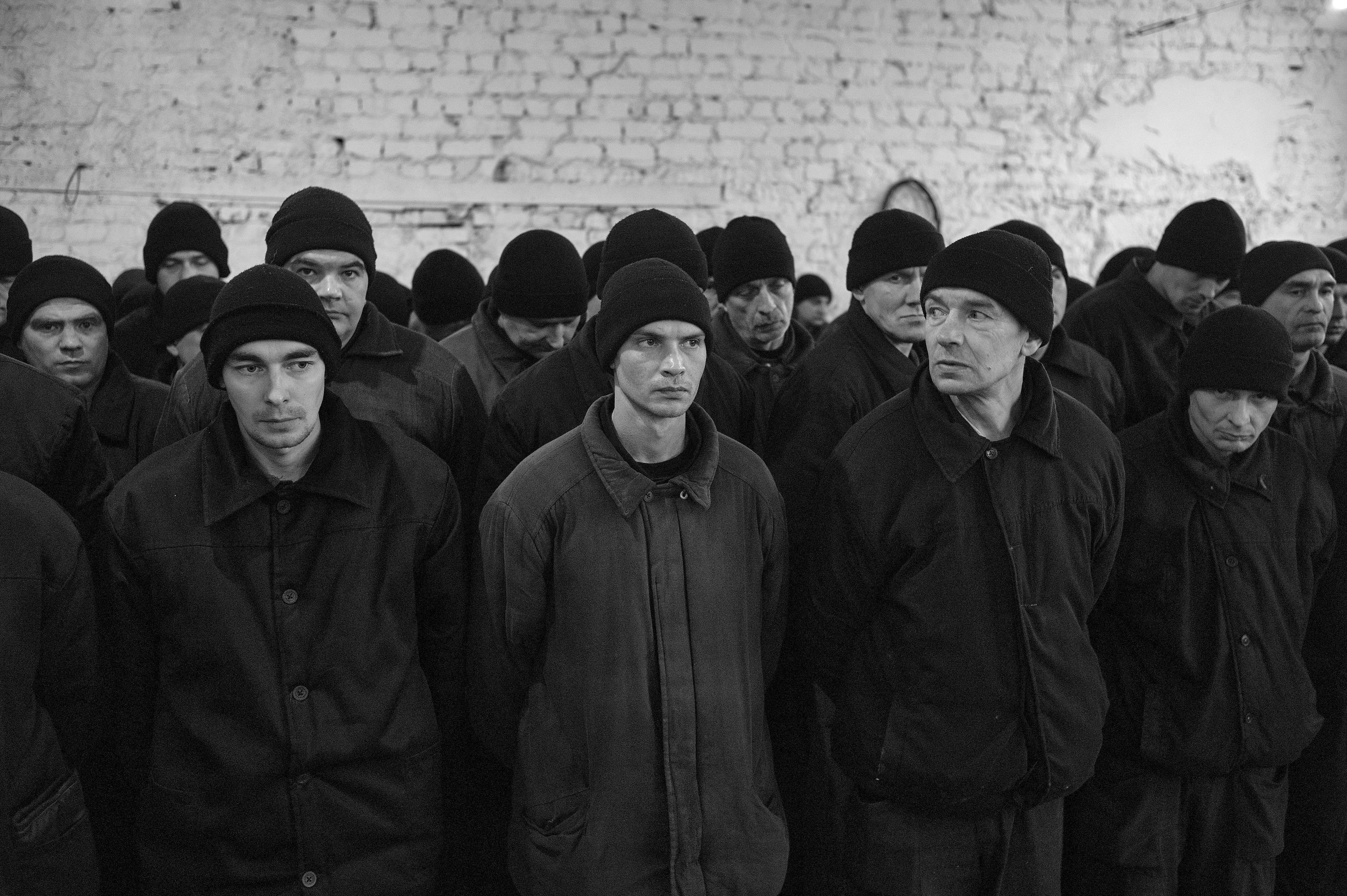 zdjecie czarno-białe. Mężczyźni w ciemnych kurtkach i czarnych czapkach stoją pod bielonym murem