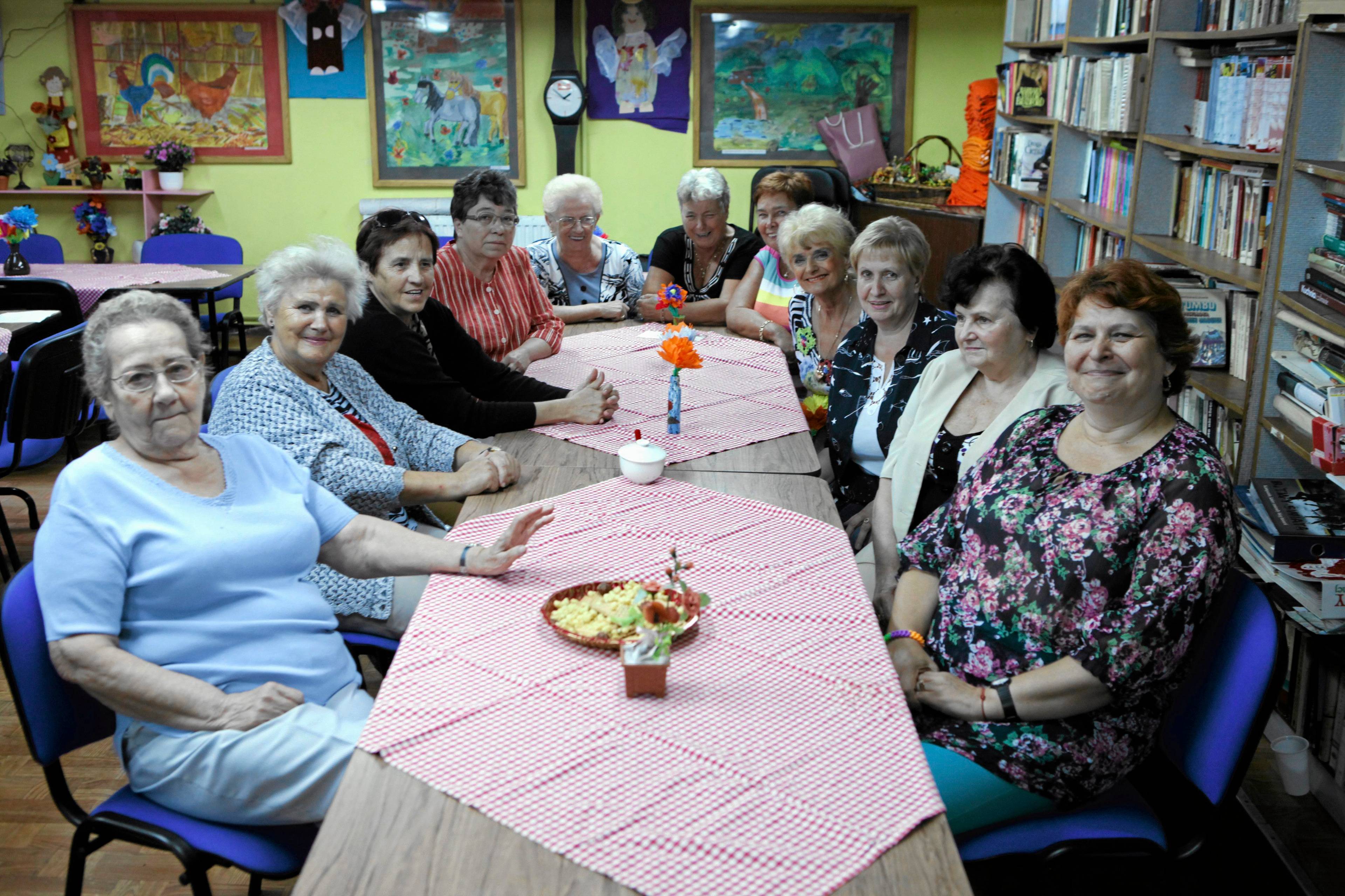 Grupa starszych kobiet siedzi wokół podłużnego stołu i patrzy w obiektyw aparatu