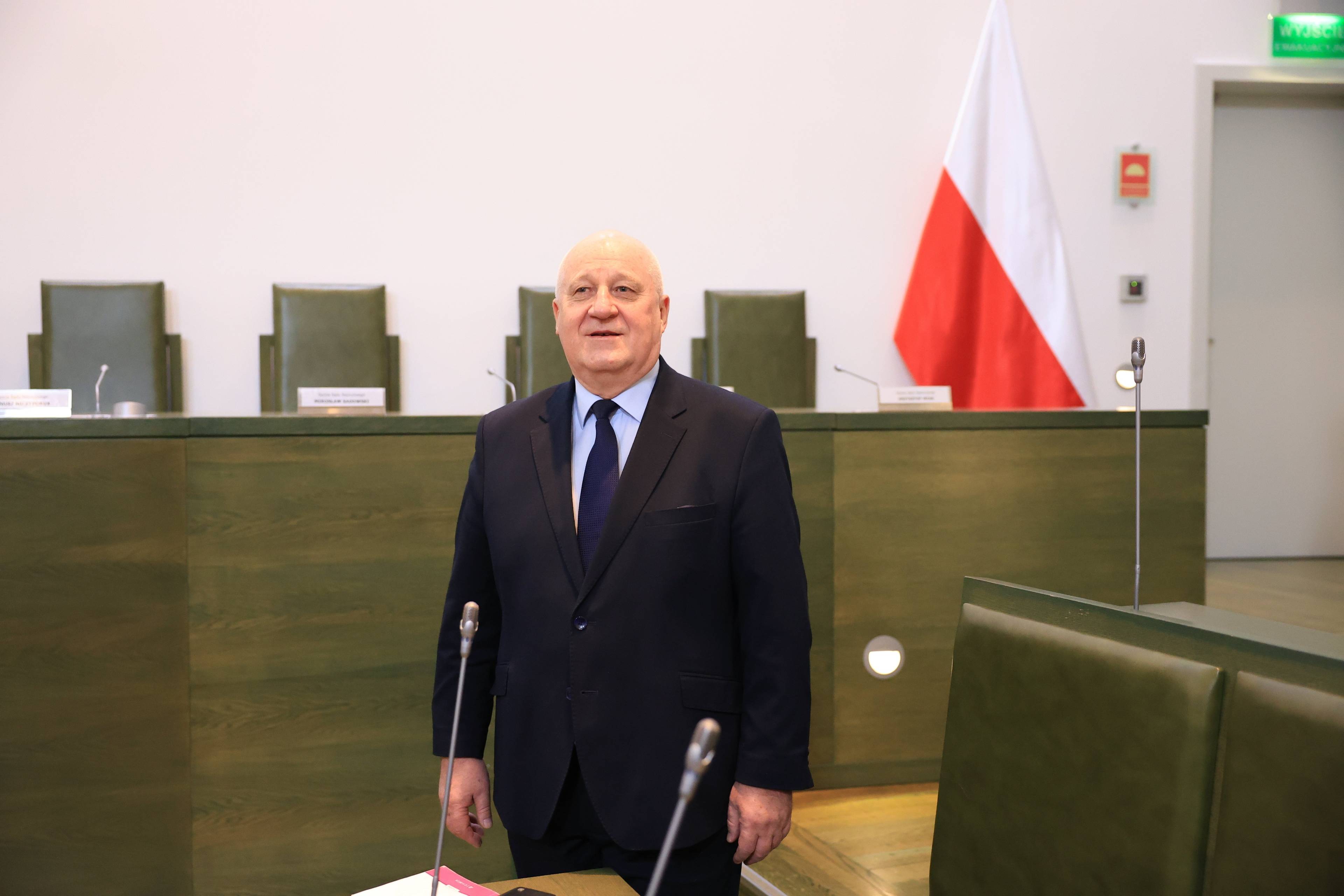 Mężczyzna w garniturze stoi na sali sądowej, za nim stół prezydialny i flaga Polski