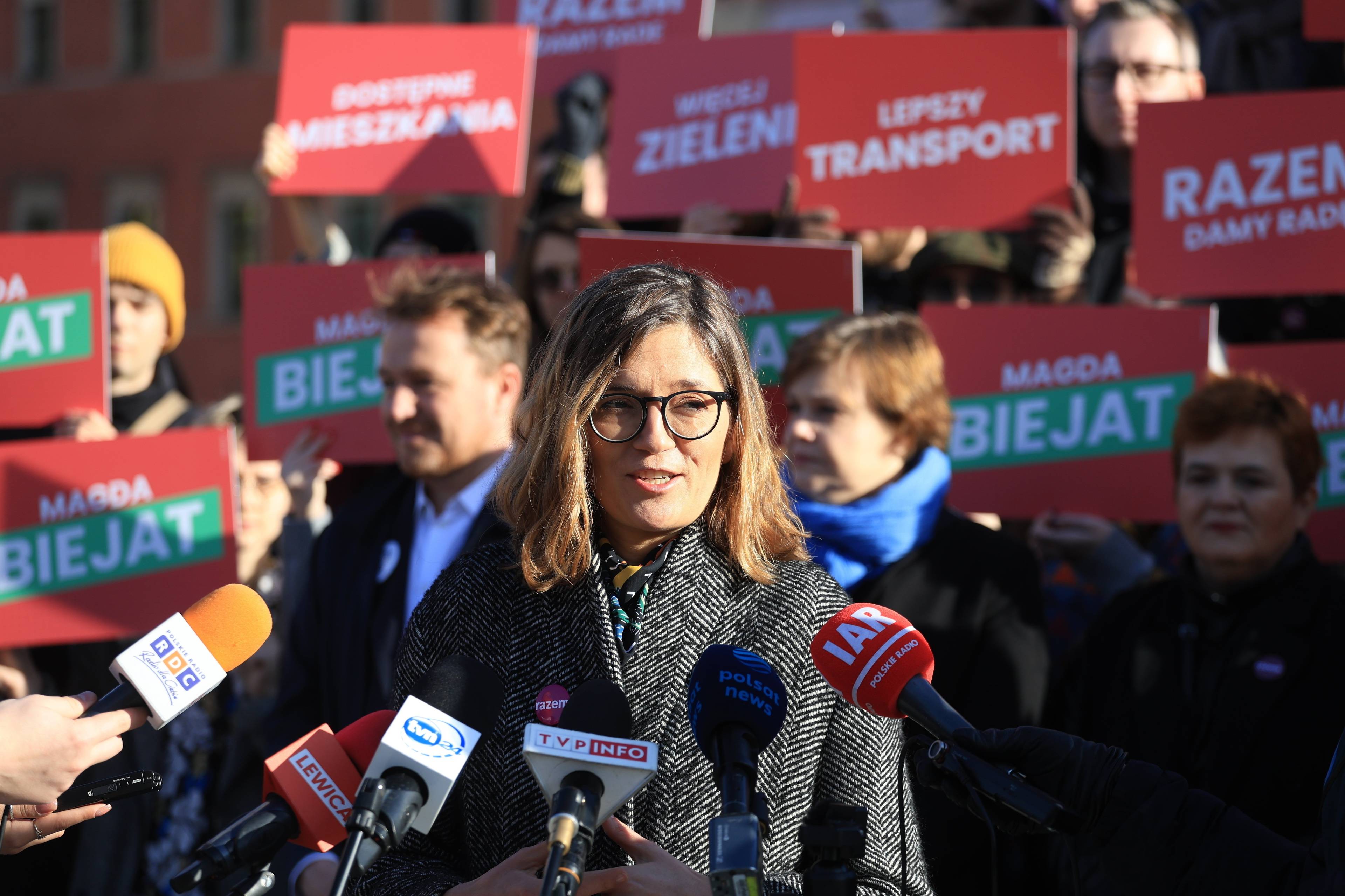 Magdalena Biejat przed mikrofonami, na tle plakatów wyborczych