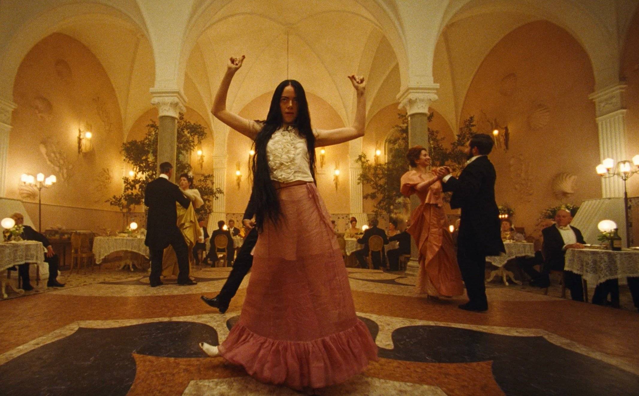 Młoda kobieta tańczy w sali balowej z uniesionymi rękoma. Emma Stone w filmie Biedne istoty