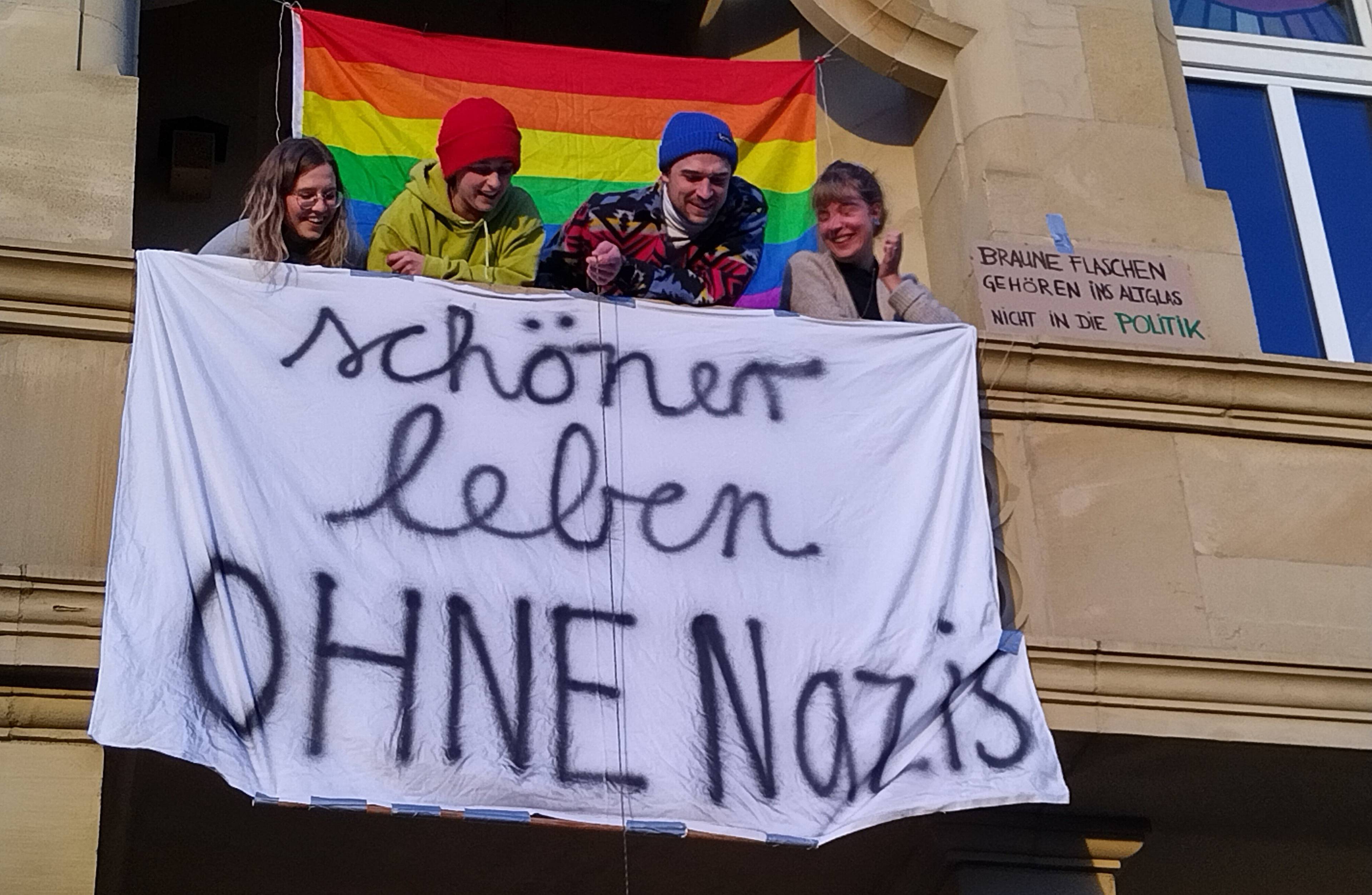 uśmiechnięci młodzi ludzie wychylają sie z balkonu i patrzą na wywieszone pod balkonem białe płótno z napisem po niemiecku: "Lepiej żyć bez nazistów". Za nimi w oknie duża tęczowa flaga.