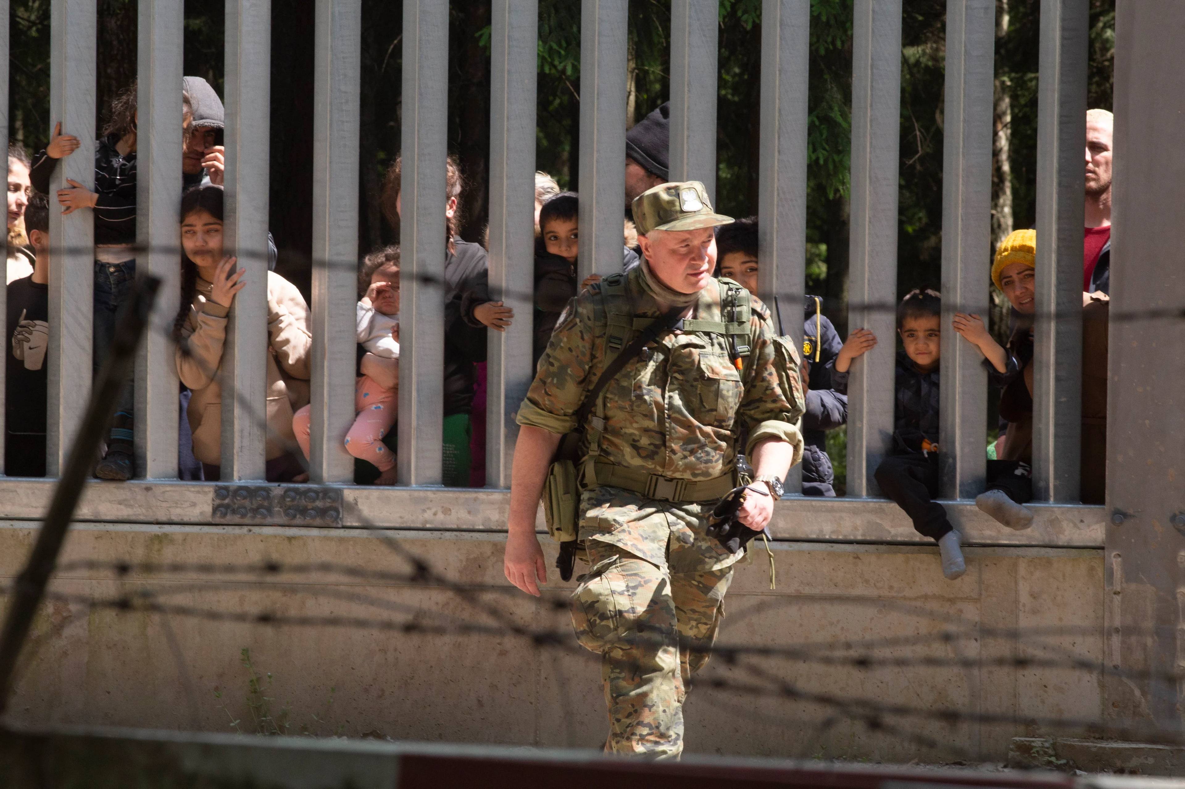 Mur na granicy, zza muru wyglądaja twarze uchodźców i migrantów