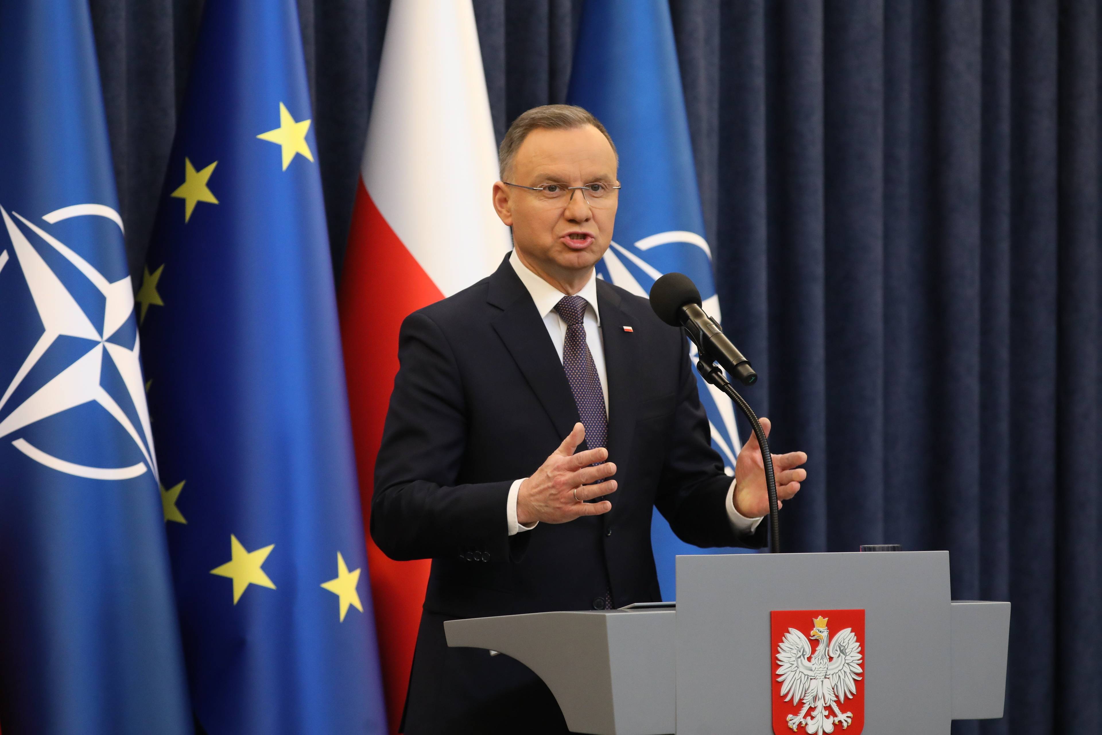 Andrzej Duda gestykuluje. Z tyłu niebieska flaga Unii Europejskiej i flaga biało-czerwona