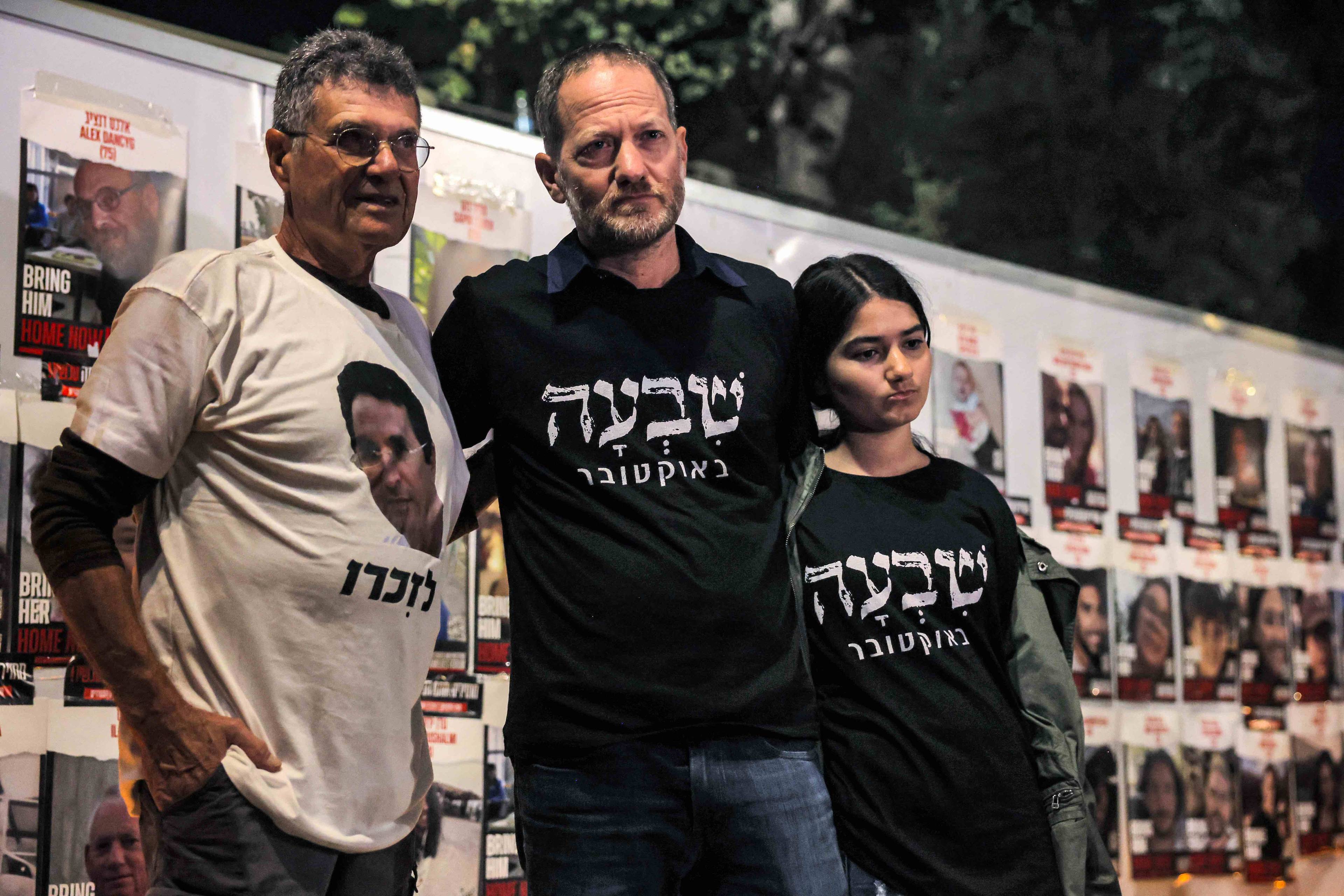 Dwóch mężczyzn w tiszertach i dziewczynka stoją na tle ściany z fotografiami, upamiętniającymi masakrę 7 października w Izraelu.