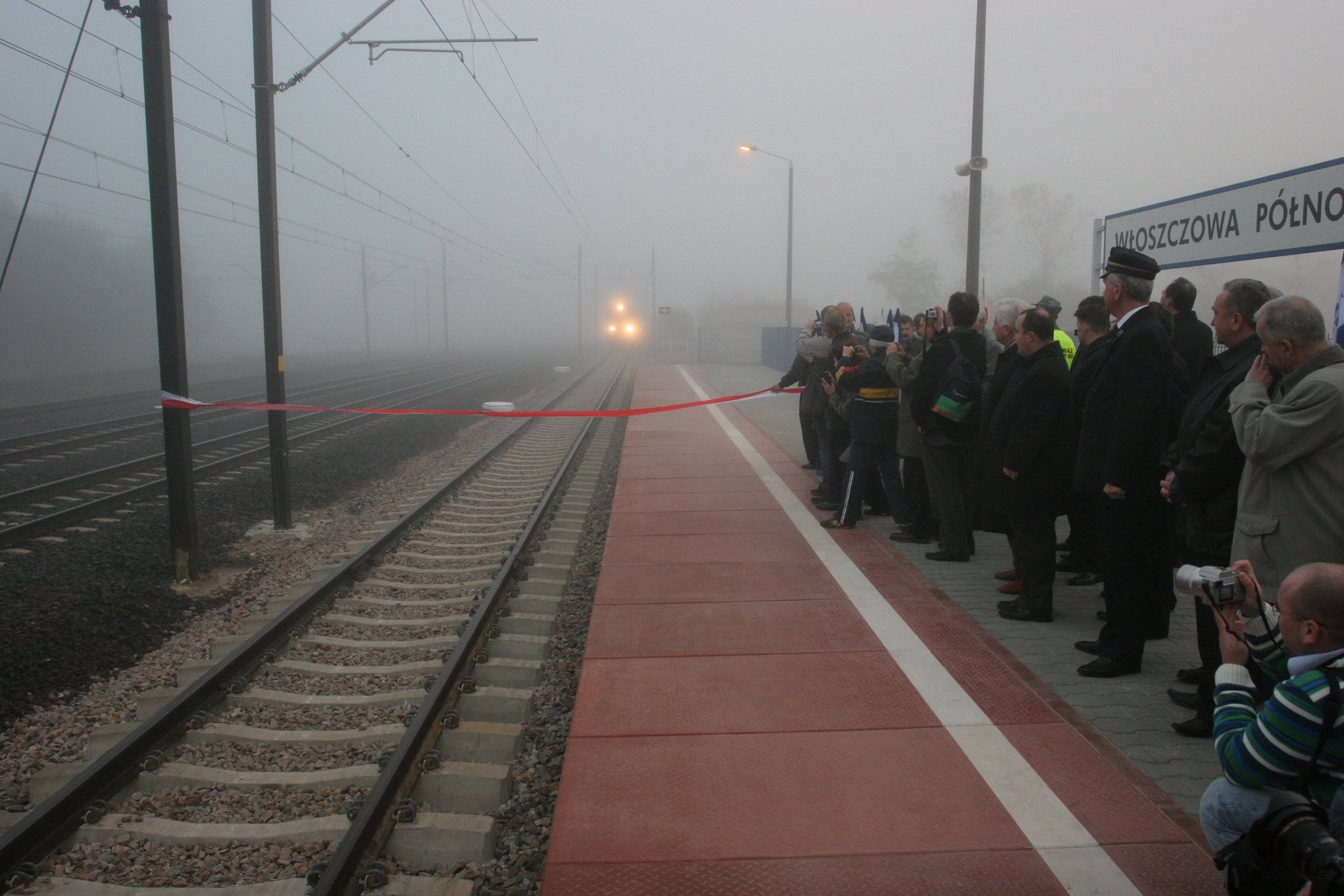 Mgła. Grupka ludzi stoi na peronie kolejowym. W oddali światła nadjeżdżającego pociągu.
