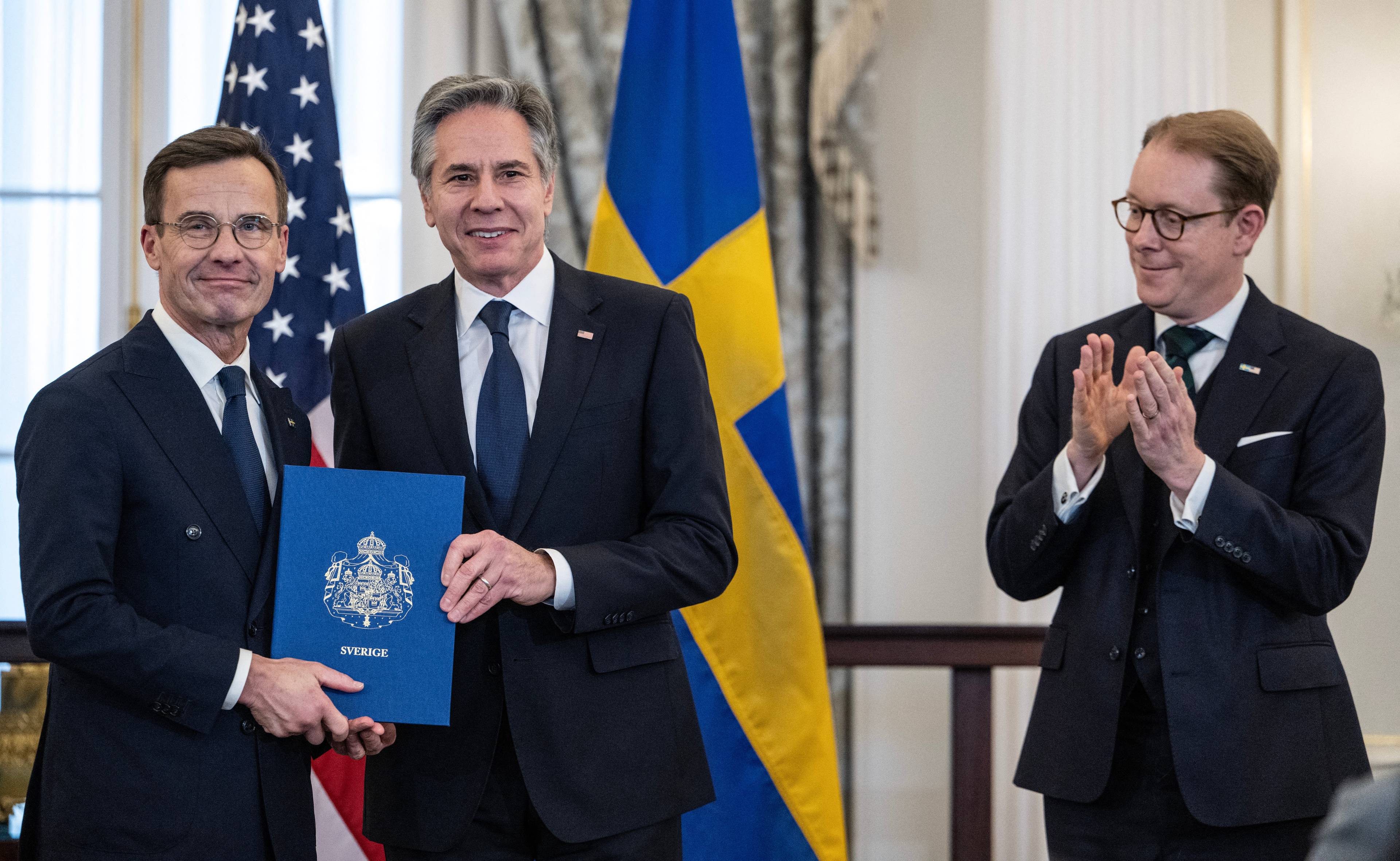 Dwóch uśmiechniętych mężczyzn w garniturach trzyma dokument w twardej, niebieskiej oprawie. Za nimi flagi Szwecji i USA