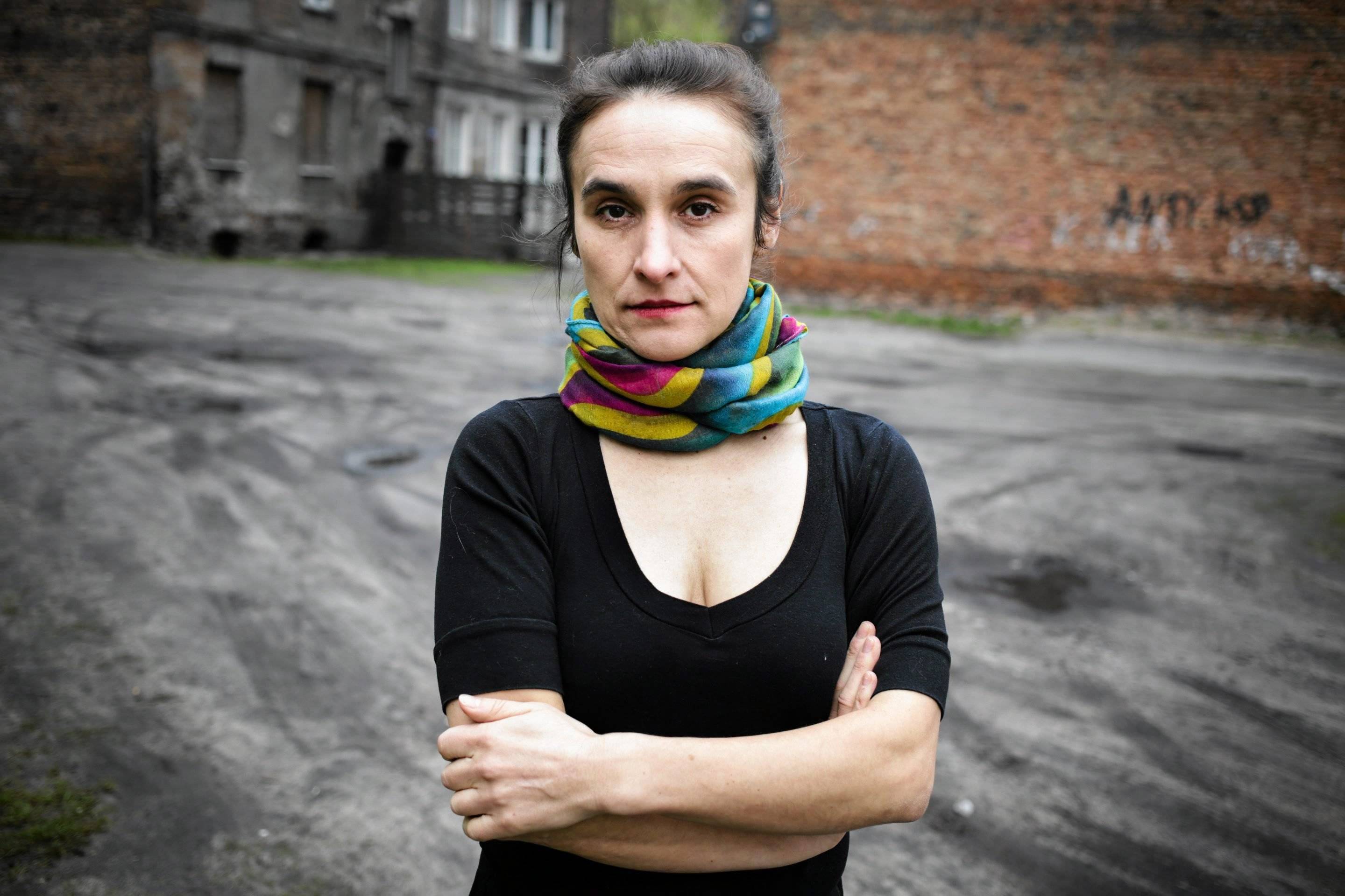 Aneta Prymaka-Oniszk. Na zaniedbanym miejskim podwórku stoi kobieta w średnim wieku, ciemne włosy zebrane do tyłu, szeroka chusta zamotana na szyi, czarna bluzka z dekoltem, ręce założone przed sobą. Mniejszość białoruska