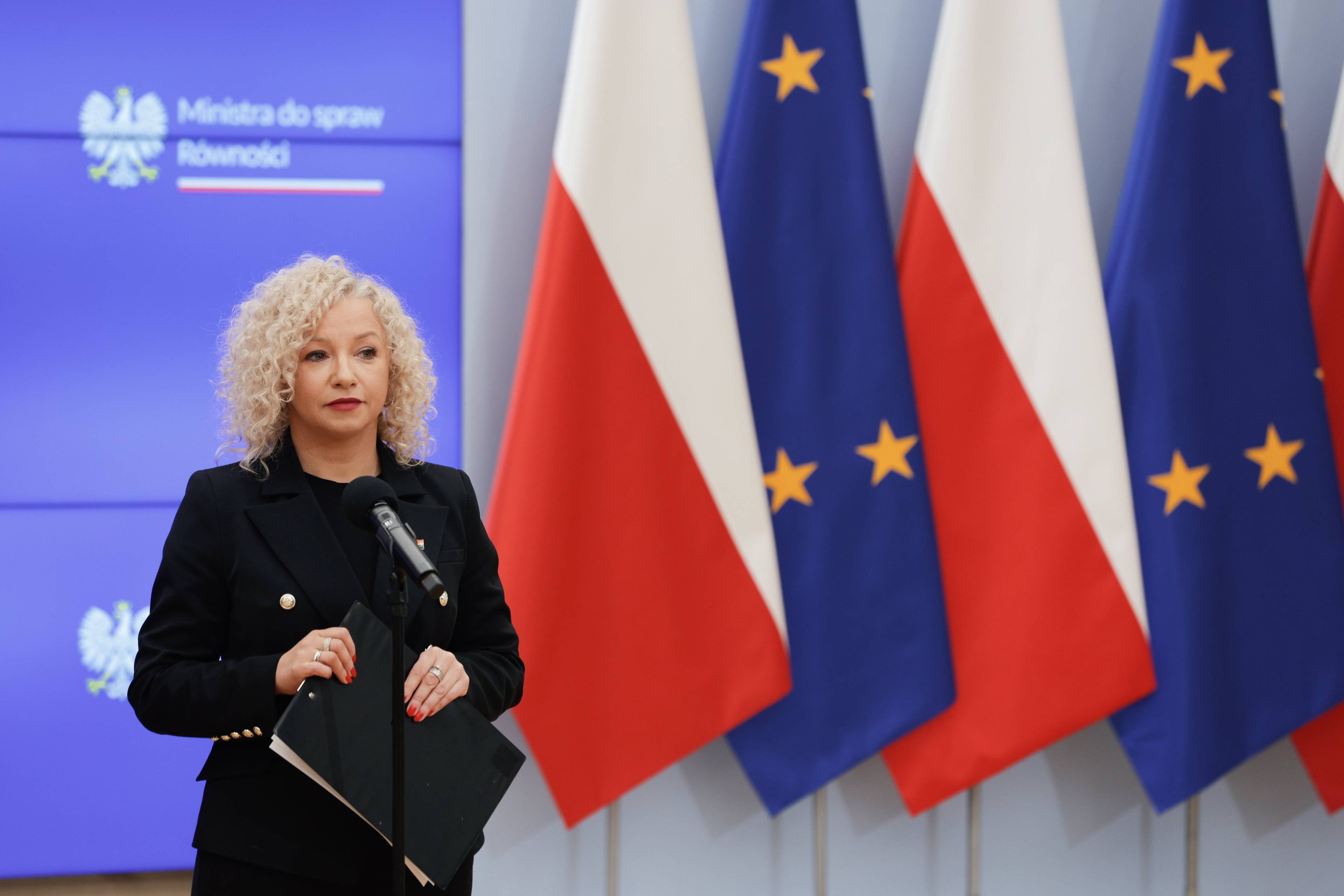 Kobieta w ciemnym żakiecie stoi na tle flag UE i Polski