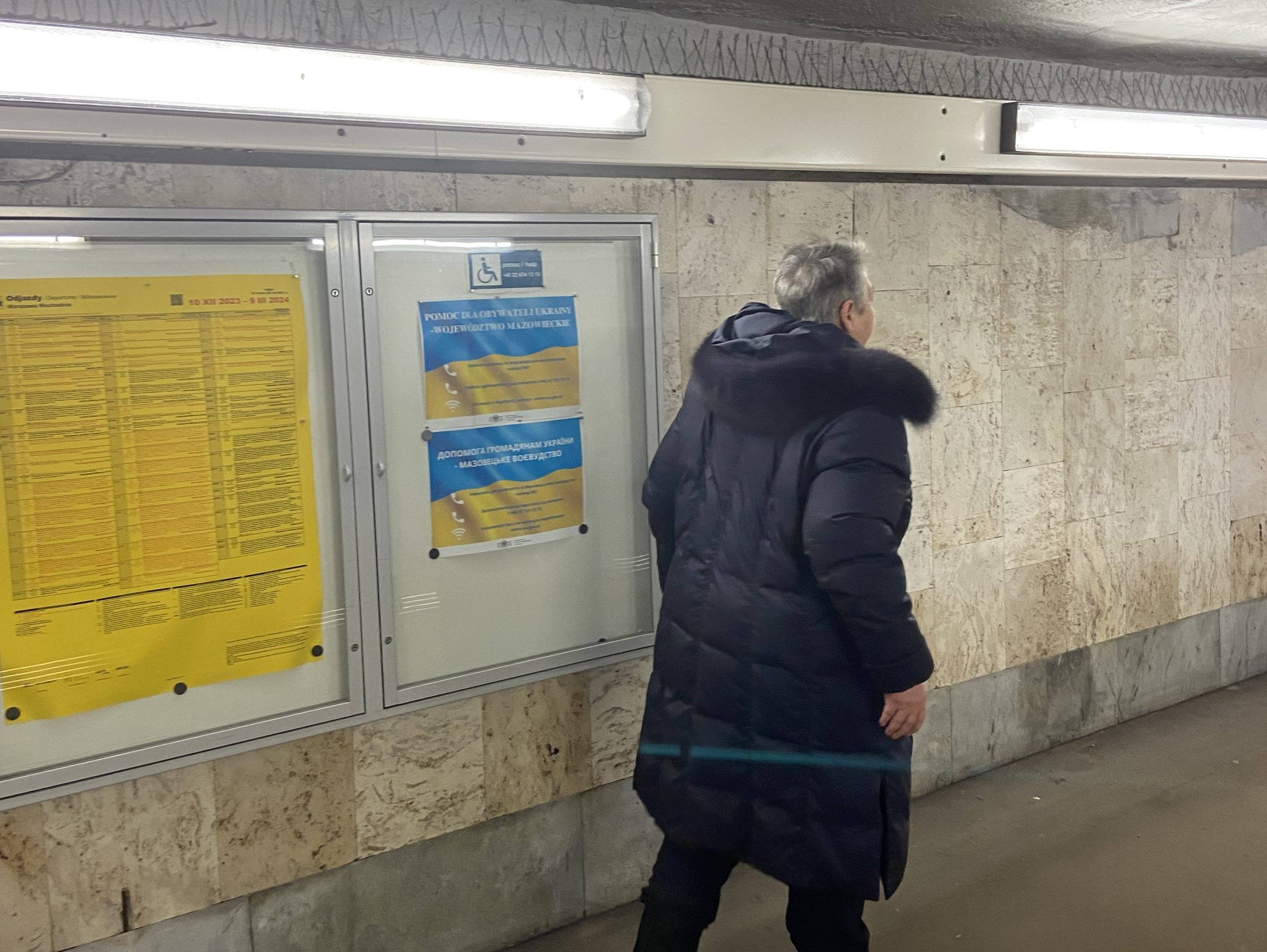 Kobieta o siwych włosach w ciemnej kurtce idzie przejściem pod peronami, na ścianie tablice informacyjne po ukraińsku
