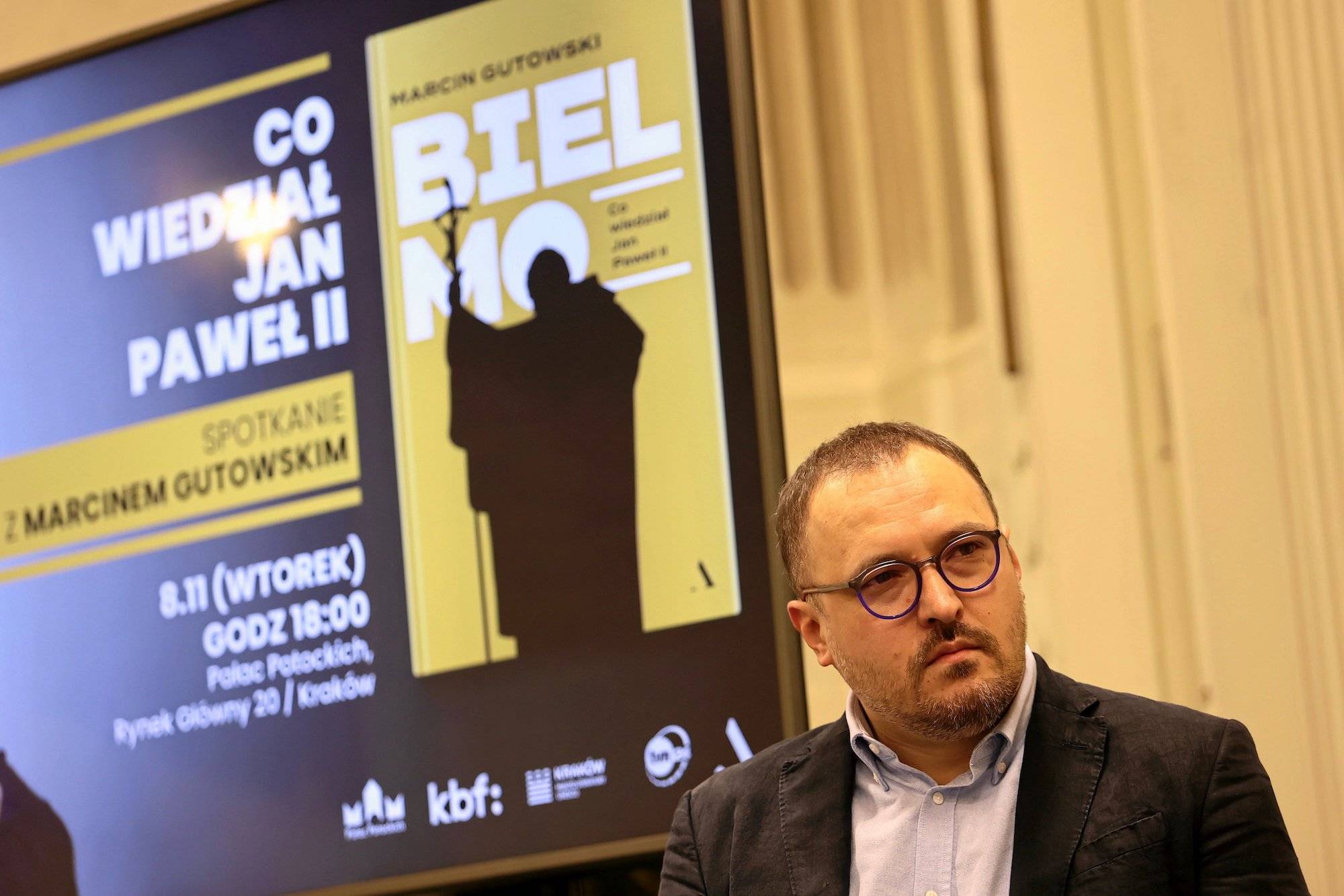 Dziennikarz Marcin Gutowski w błękitnej koszuli i ciemnej marynarce. Na twarzy ma okrągłe ciemne okulary. W tle plakat z okładką jego książki "Bielmo"