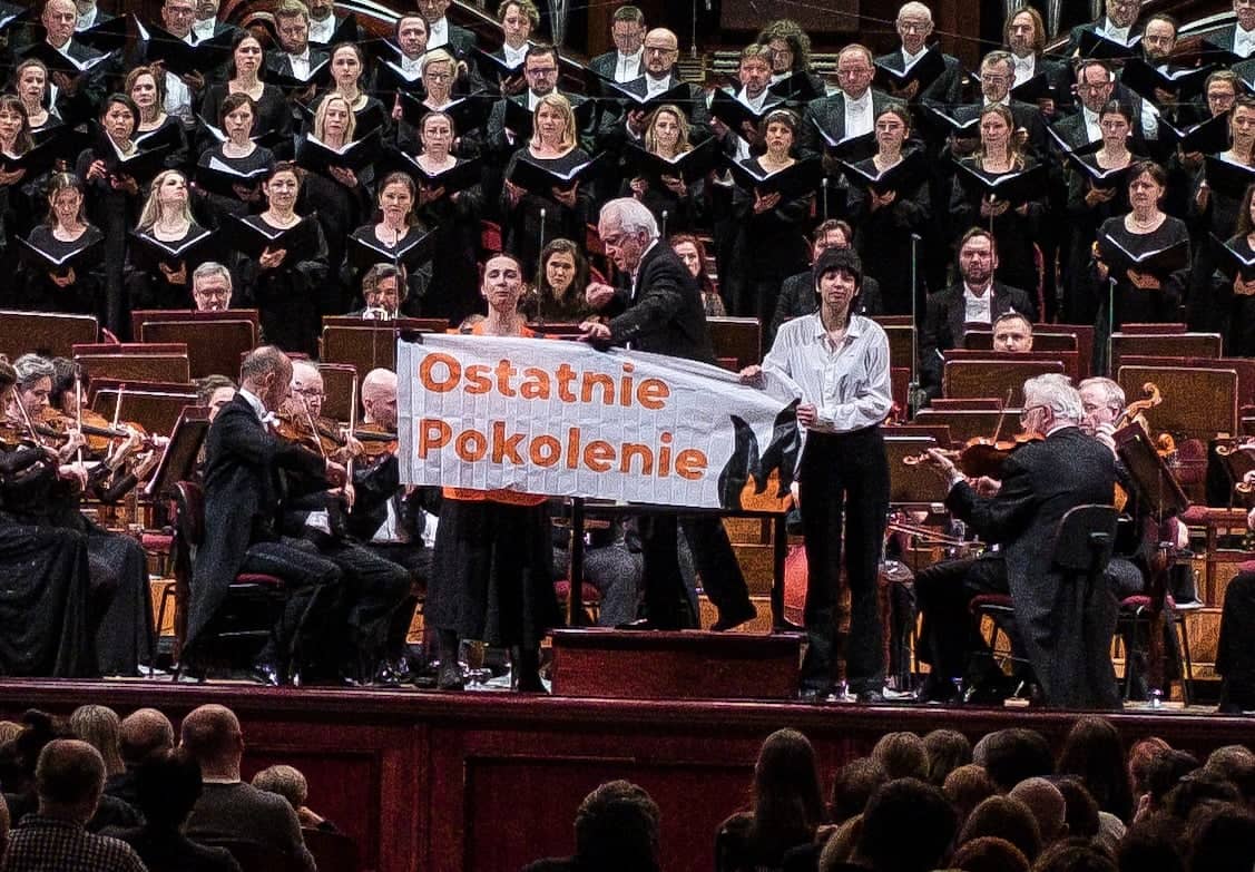 Dwie aktywistki stoją na scenie filharmonii podczas koncertu, trzymając transparent z napisem „Ostatnie Pokolenie” i rysunkiem płomienia.