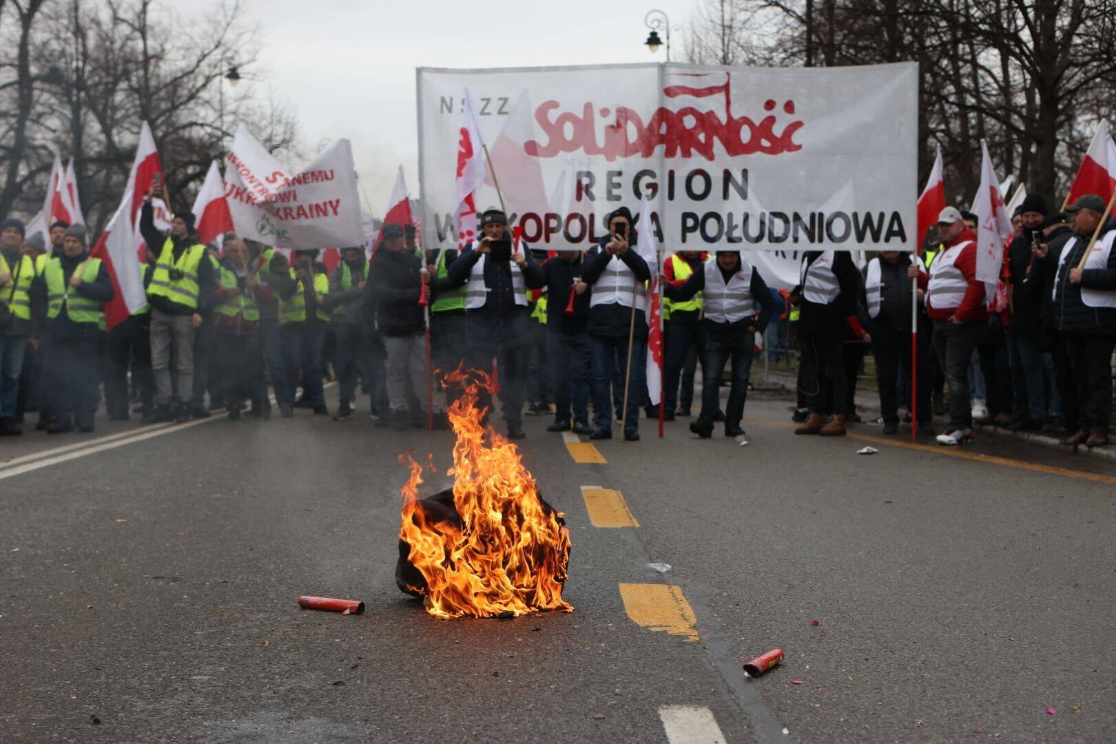 Na pierwszym planie palona opona, w tle grupa protestujących rolników w żółtych kamizelkach, z polskimi flagami.