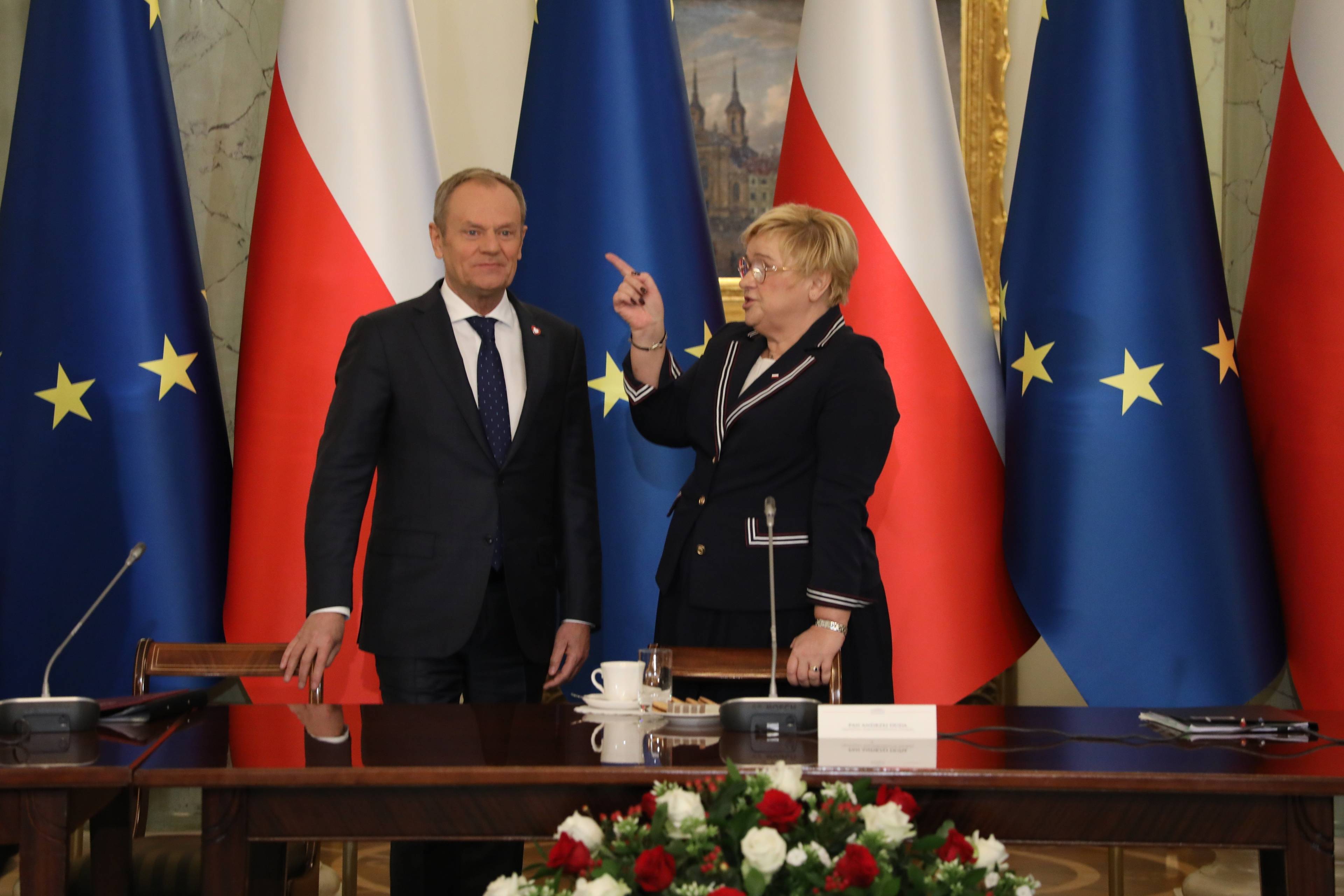 Kobieta w ciemnej garsonce (po prawej) wskazuje palcem na Donalda Tuska (po lewej). W tle flagi polski i Unii Europejskiej