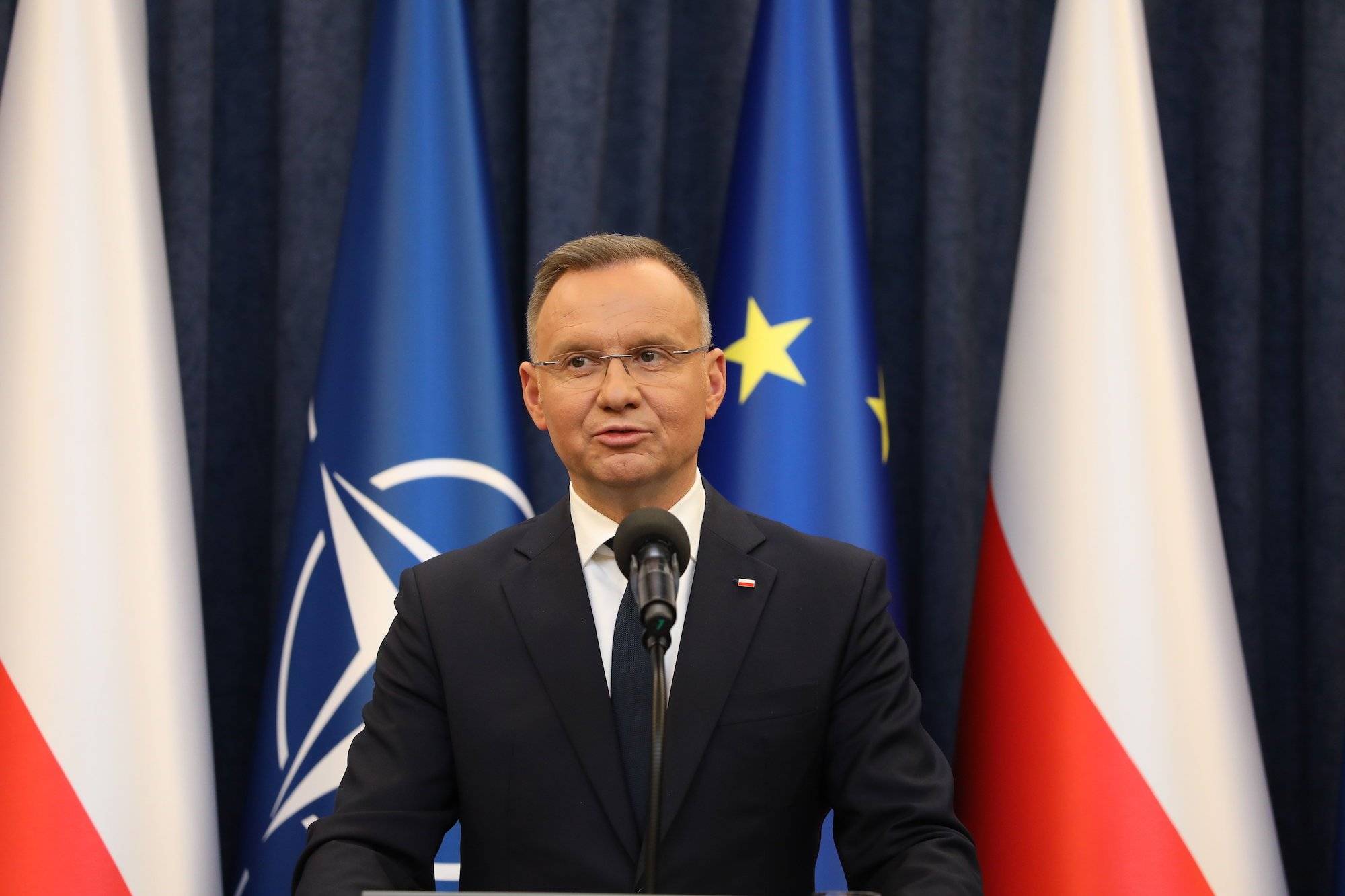 Prezydent Andrzej Duda na tle flag UE, Polski i NATO. Ubrany w ciemny garnitur, na twarzy okulary. Przemawia do mikrofonu
