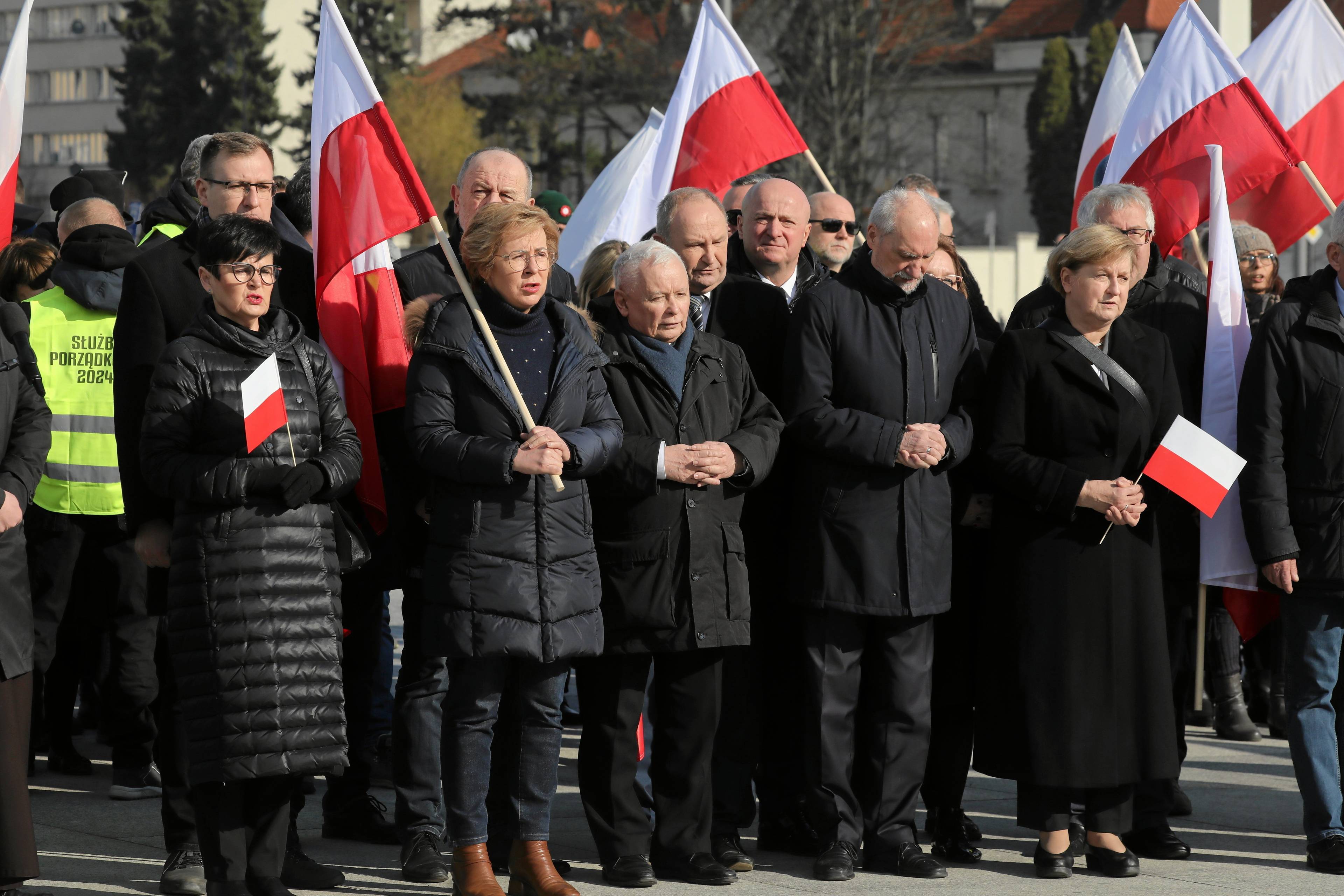 Ludzie z białoczerwonymi flagami i w czarnych kurtkach. W środku - Jarosław Kaczyński