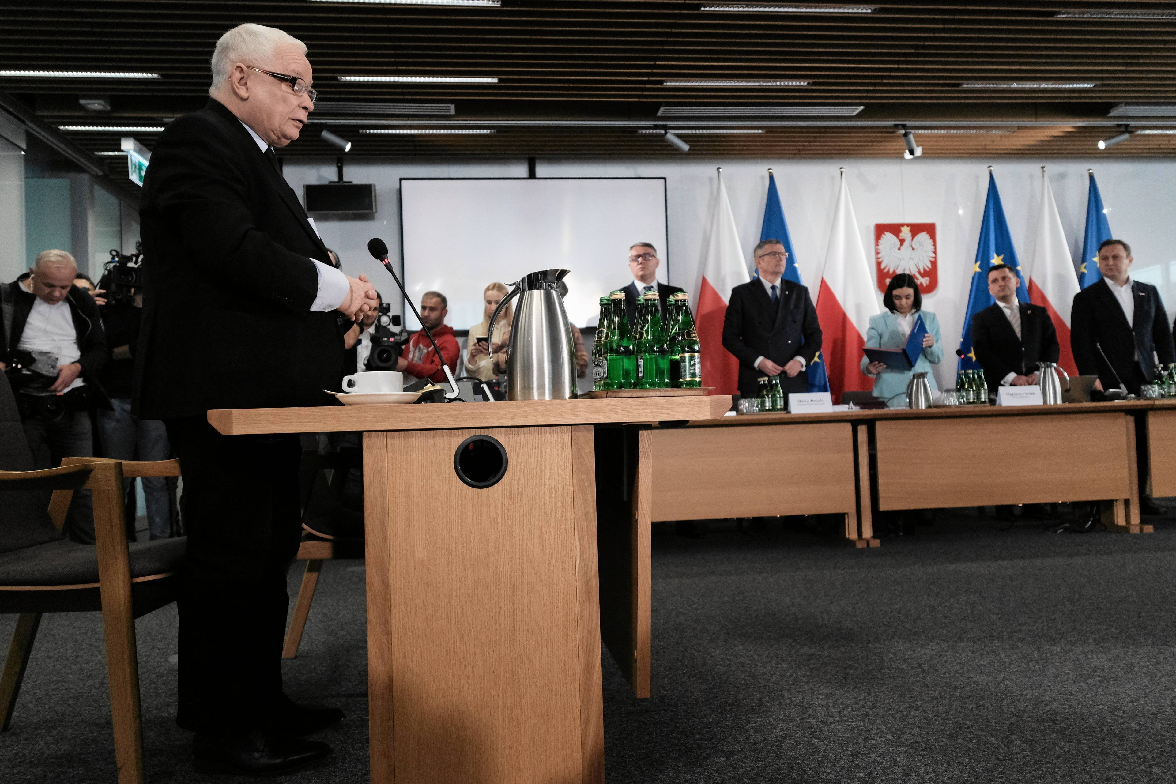Jarosław Kaczyński stoi przy mikrofonie, za nim członkowie komisji śledczej