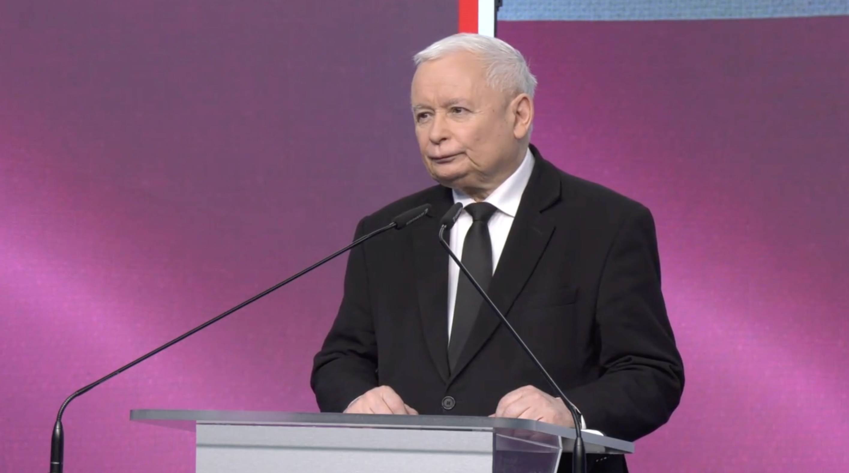 Jarosław Kaczyński przemawia podczas konferencji. Za nim różowo-stalowe tło. Kaczyński ma na sobie biała koszulę i cxarny garnitur i ciemny krawat.