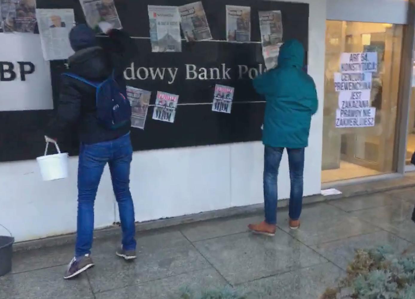 Dwie osoby przyklejającą plakaty na tablicy z napisem „NBP. Narodowy Bank Polski”. Hasło na jednym z plakatów brzmi: „Art. 54 Konstytucji: Cenzura prewencyjna jest zakazana. Prawdy nie zakneblujesz”.