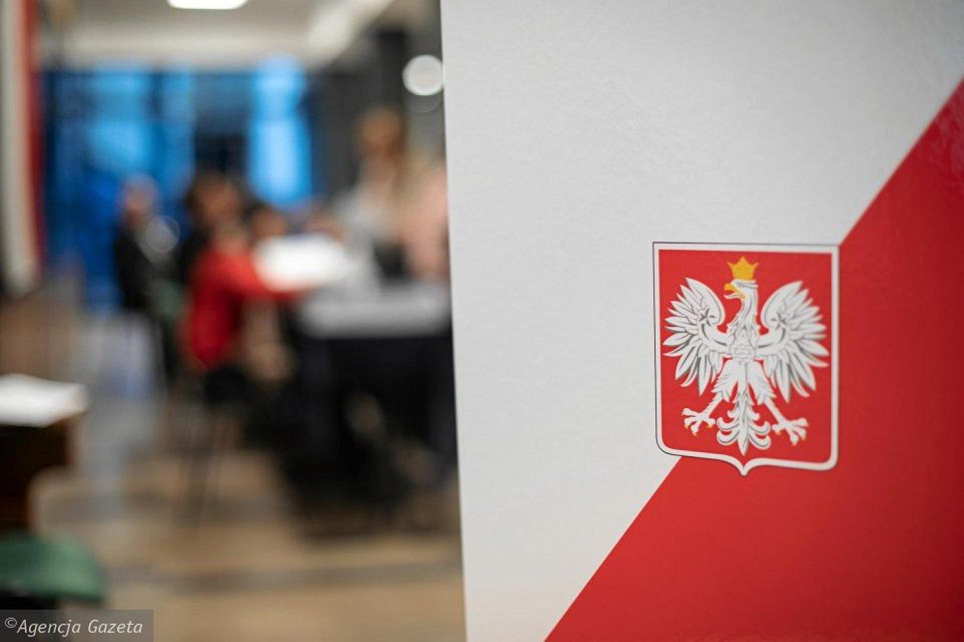Urna wyborcza w biało-czerwonym kolorze, na pierwszym planie biały orzeł, godło Polski, w tle rozmazany obraz lokalu wyborczego