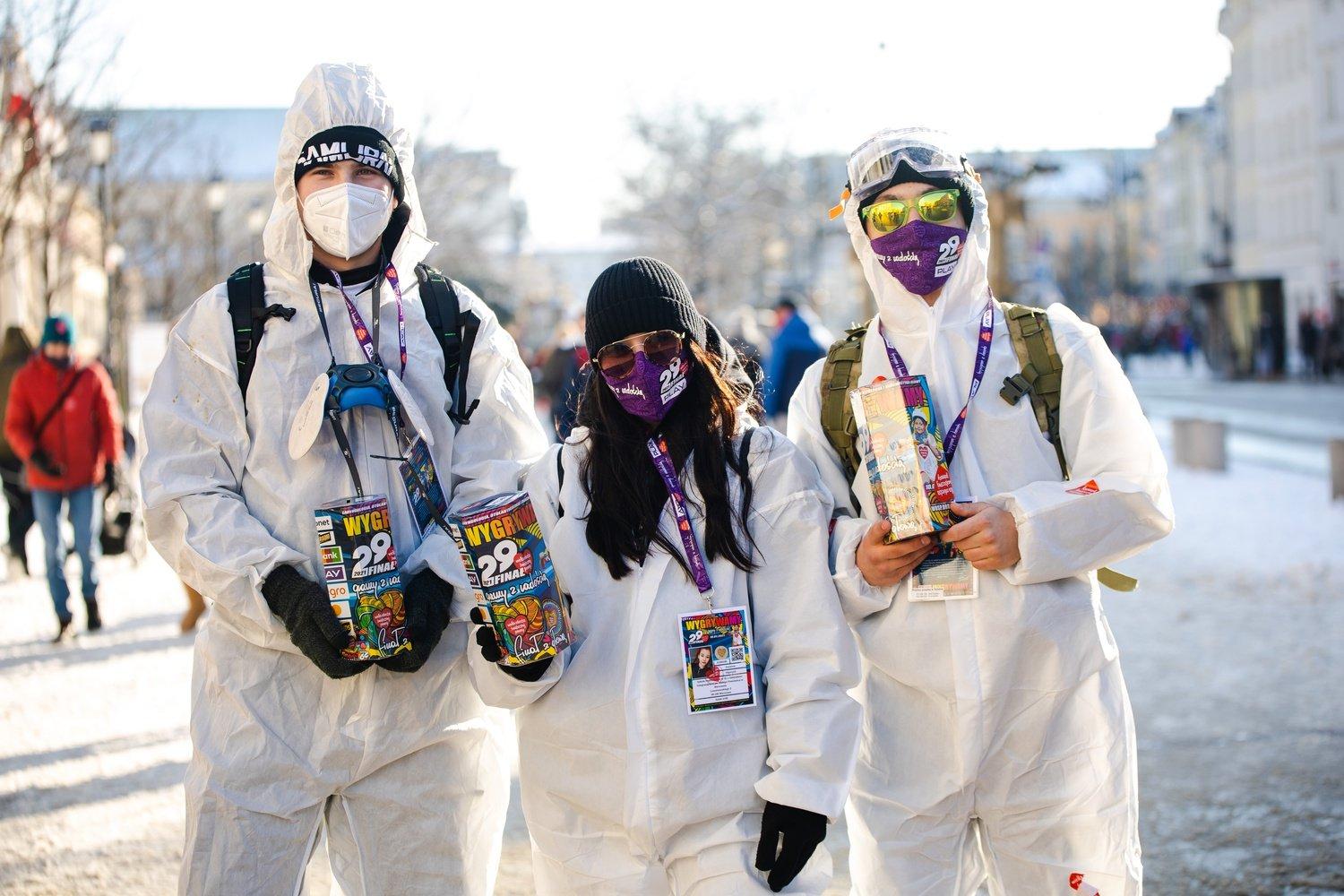 Trójka młodych wolontariuszy w białych skafandrach a białych maseczkach na twarzy z puszkami na pieniądze w rękach