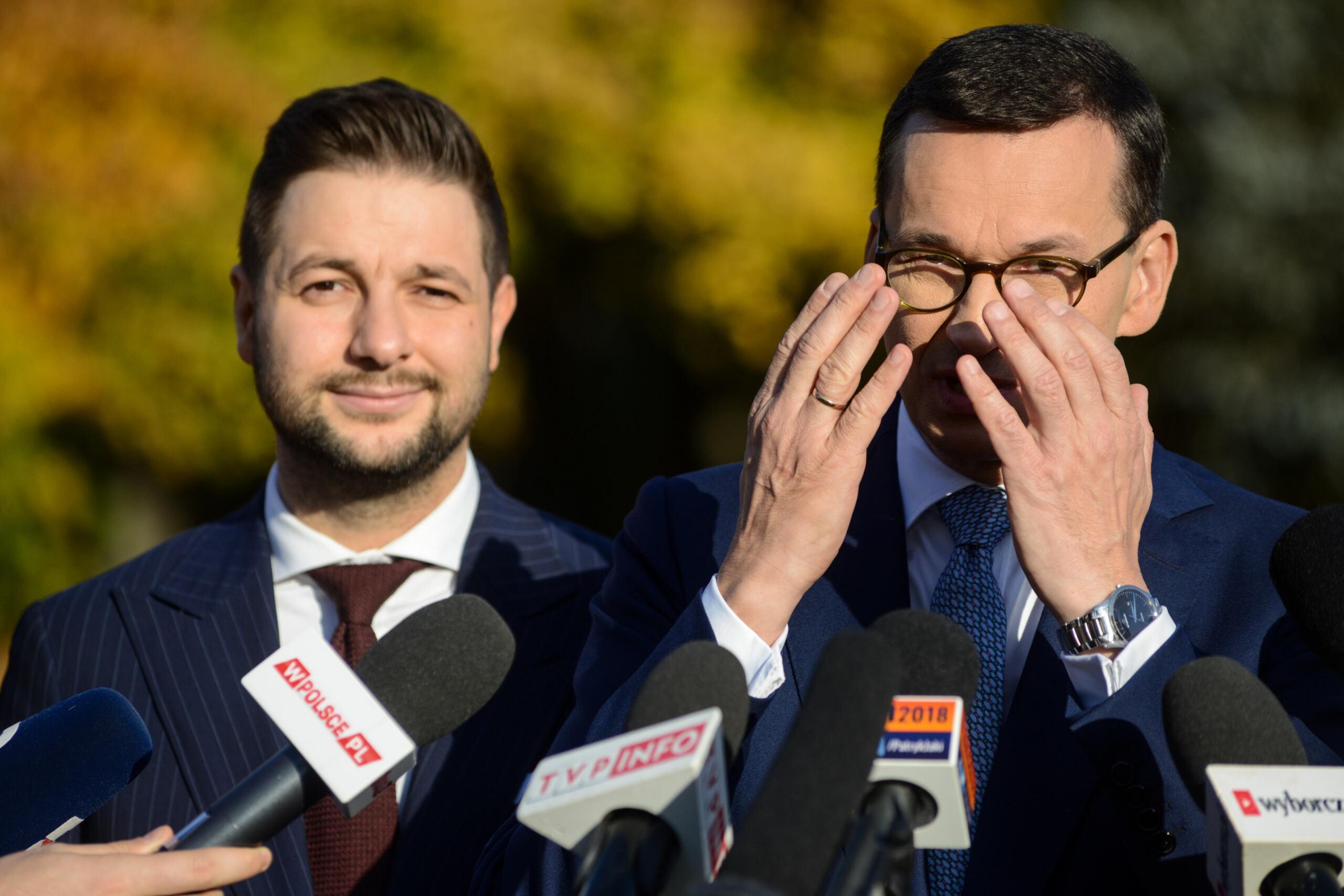 Patryk Jaki stoi obok premiera Morawieckiego, który zasłania dłońmi twarz