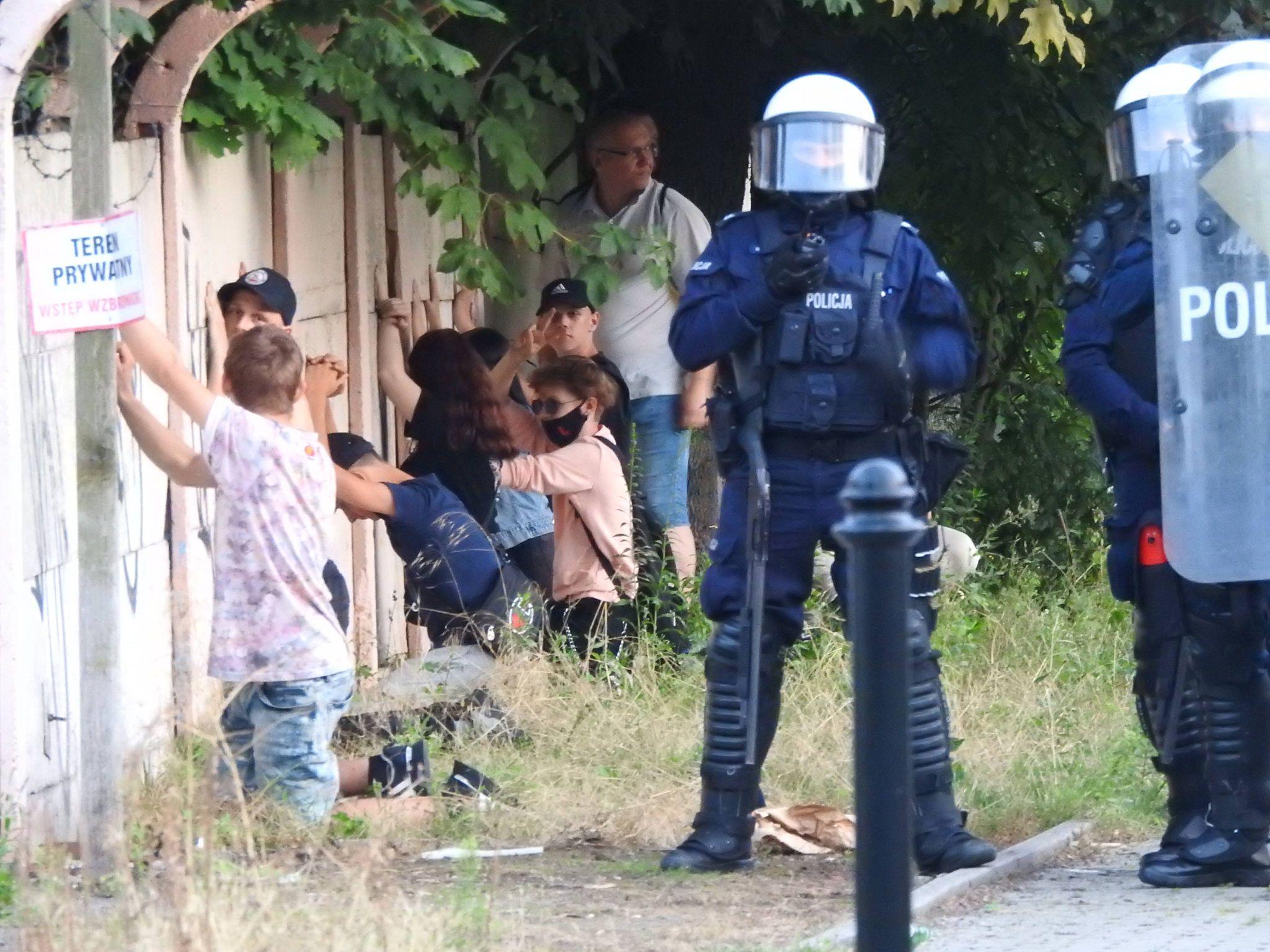 Nastolatki klęczące pod płotem z drutem kolczastym, pilnowane przez uzbrojonych policjantów