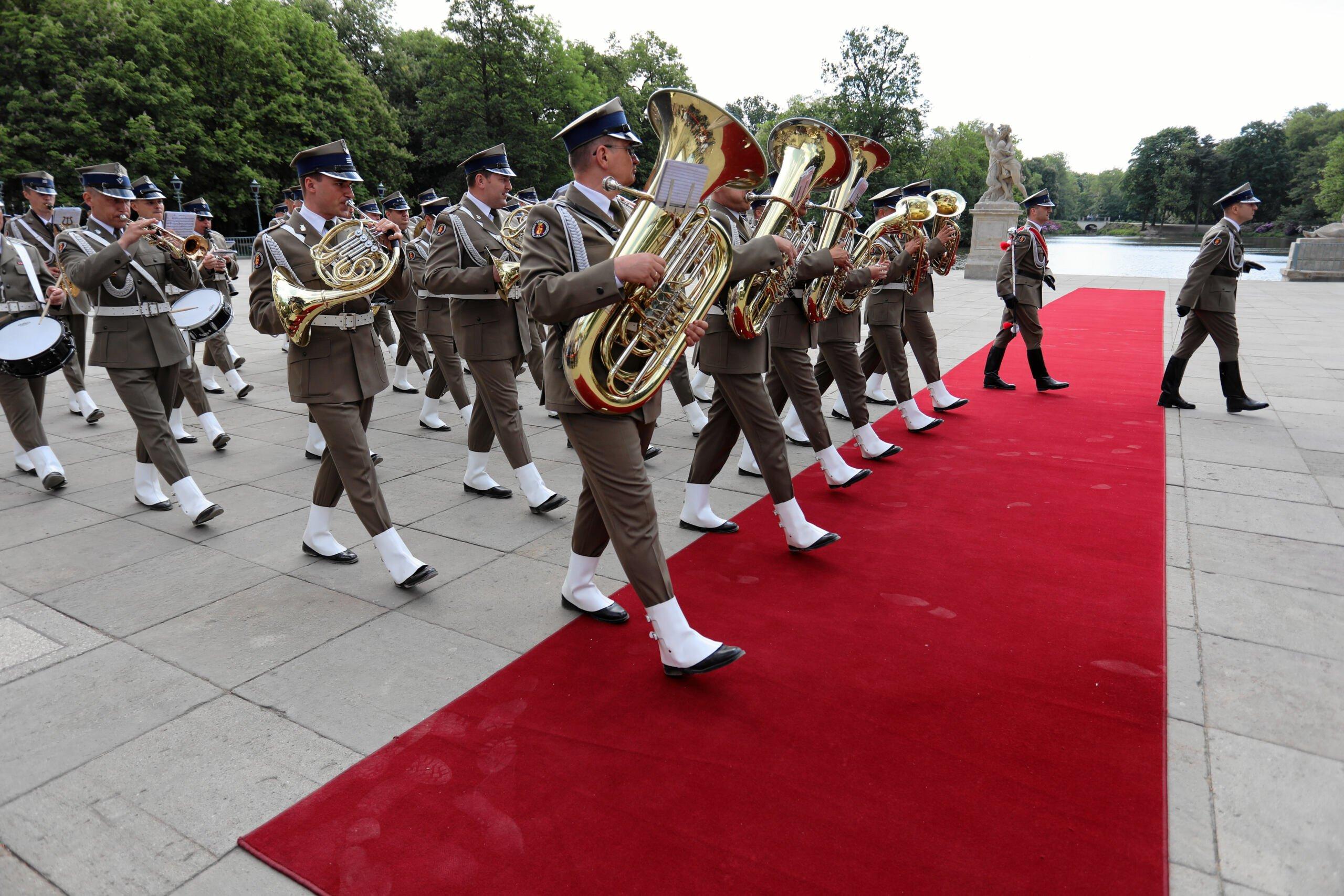 Żołnierze z orkiestry wojskowej wchodzą na czerwony dywan marszowym krokiem
