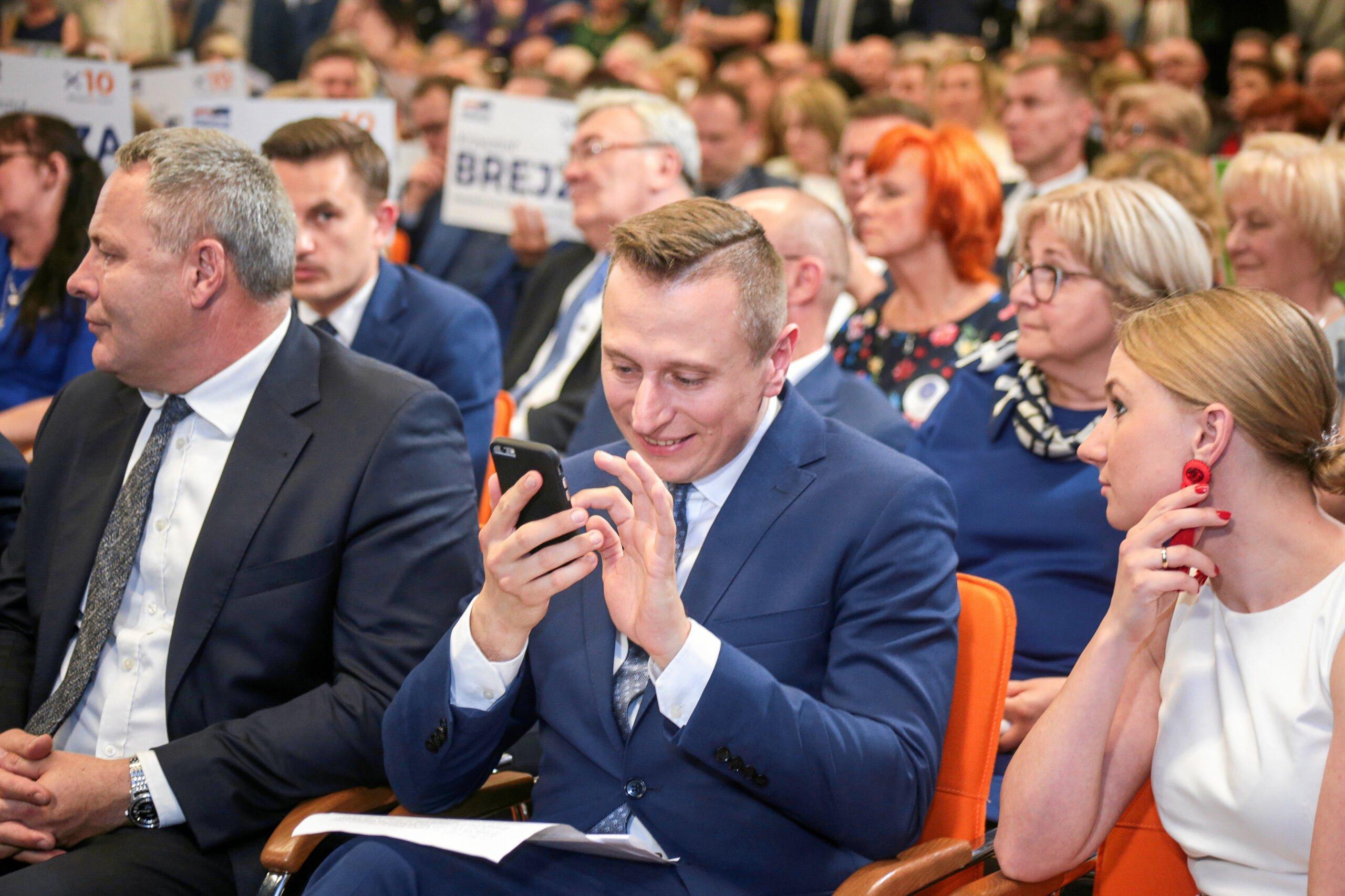Krzysztof Brejza podczas kampanii wyborczej 2019 siedzi w pierwszym rządzie i przegląda smartfona