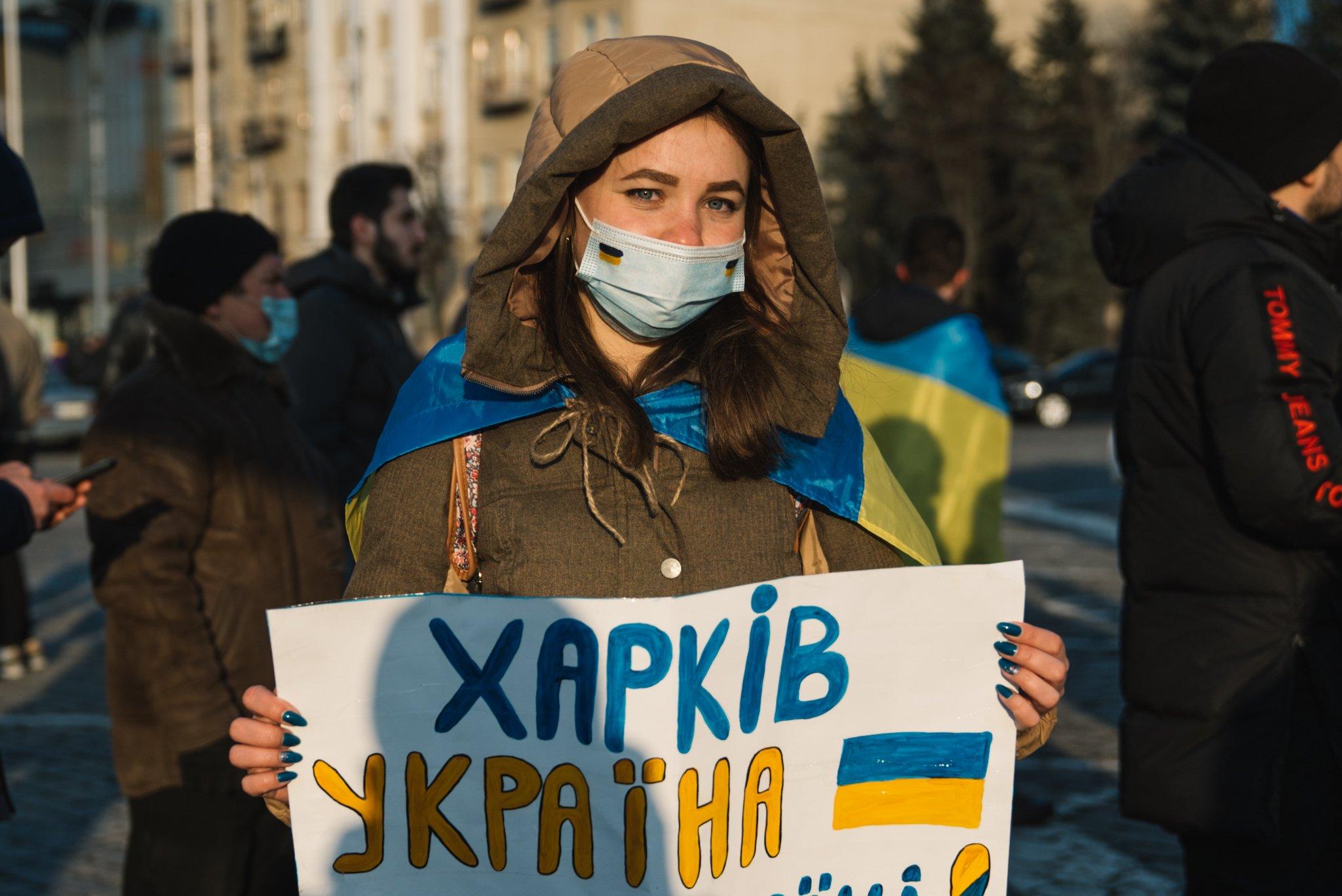 Ołesia Dekusz w zimowej kurtce i maseczce trzyma plakat "Charkiv Ukraina"