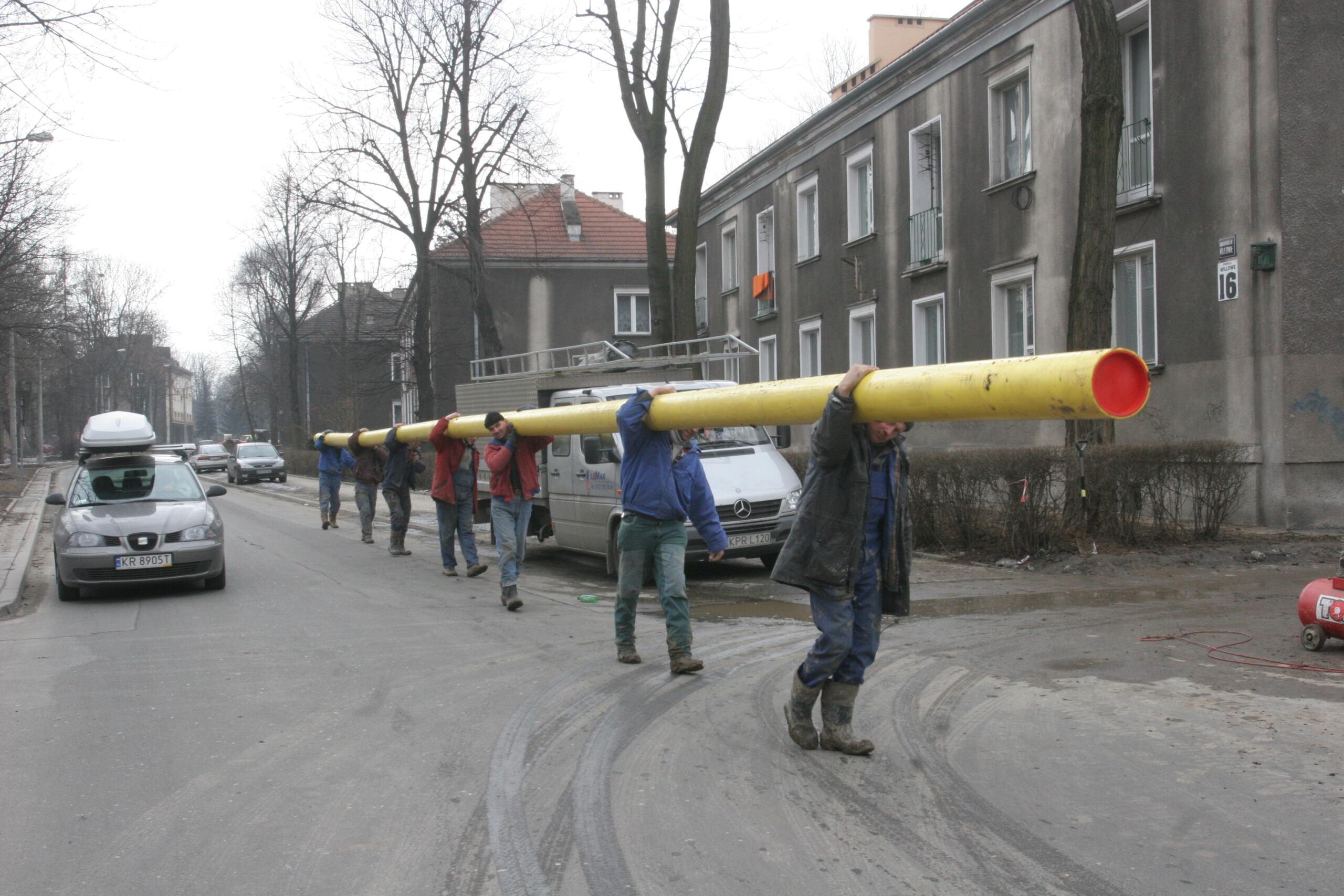 Mężczyźni niosą ulicą żóltą rurę