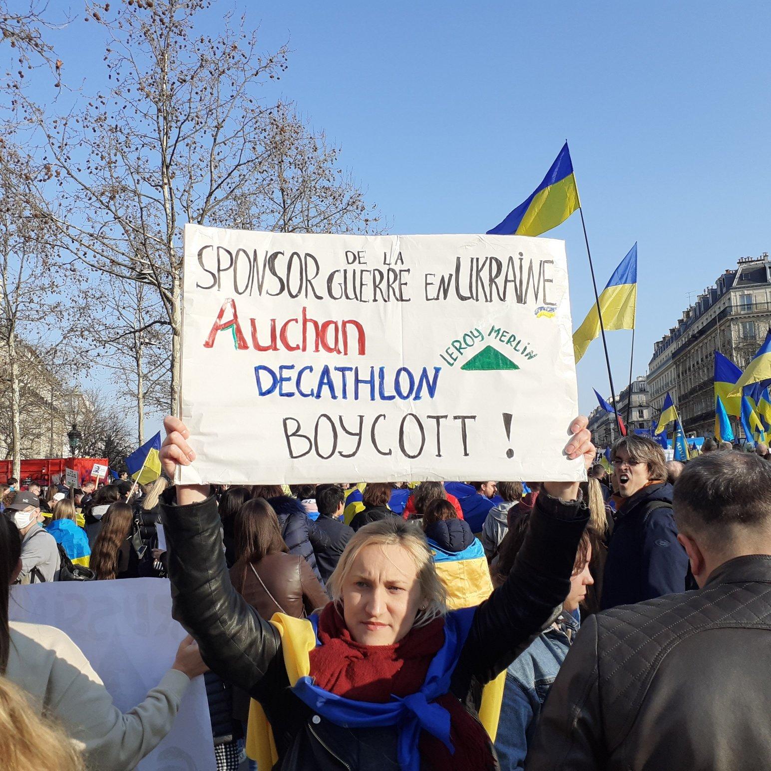 Demonstrant trzyma napis po francusku "Sponsorzy wojny w Ukrainie" pod którym narysowane są loga Auchan, Decathlon i Leroi Merlin, pod spodem dużymi literami słowo Bojkot