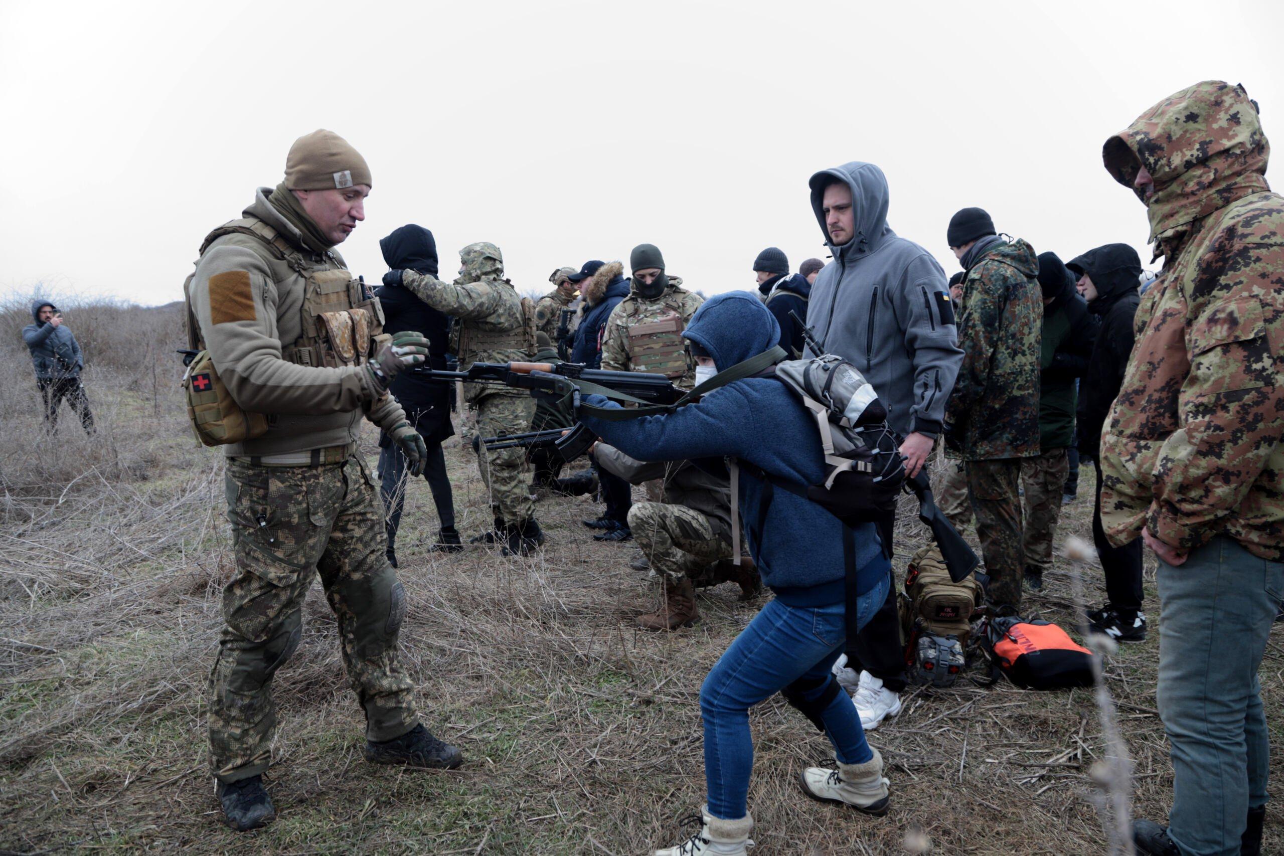 Grupa ludzi, część ubrana w wojskowe mundury, na pierwszym planie osoba w cywilnych ubraniach celuje z karabinu