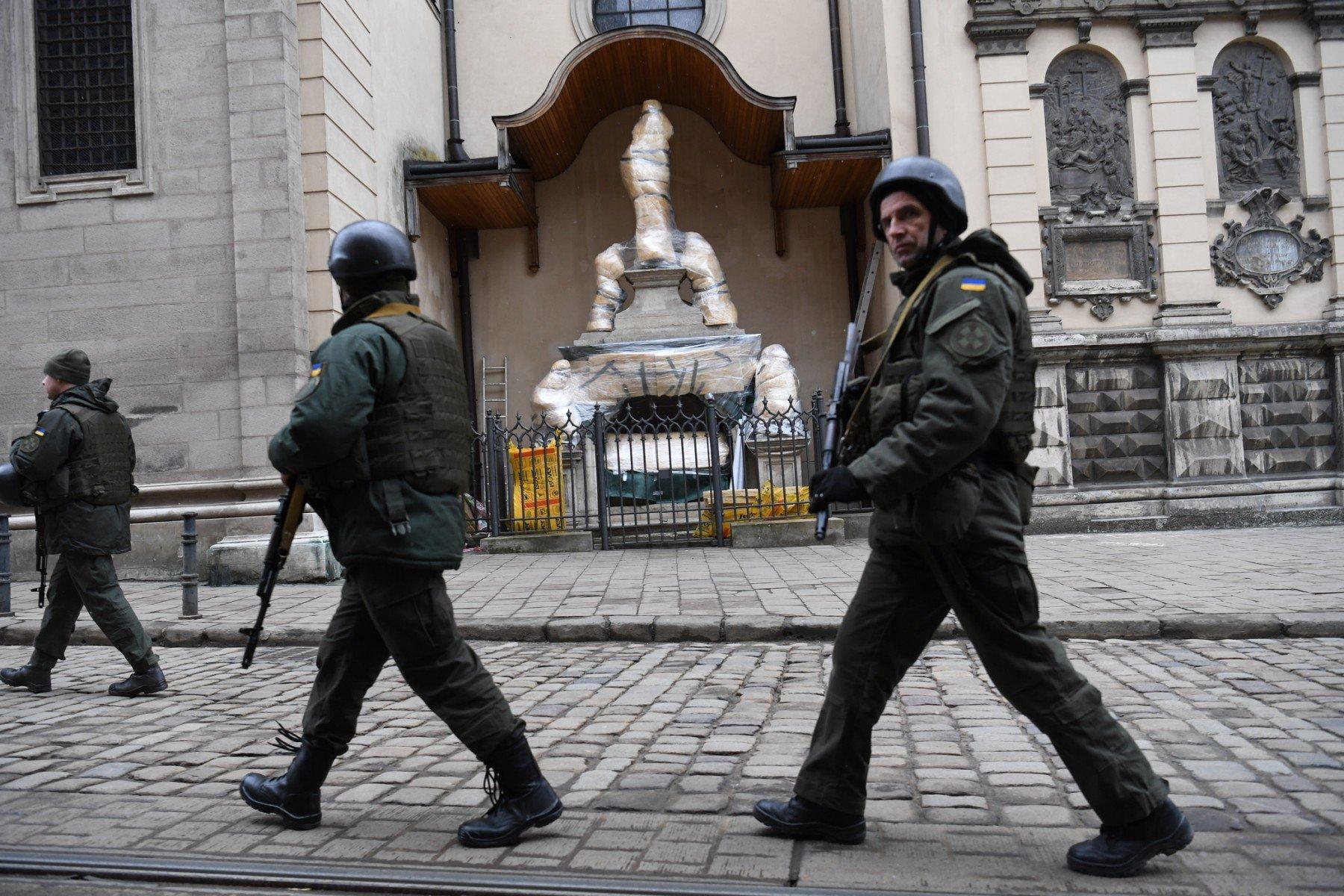 Żołnierze w pełnym uzbrojeniu na ulicy Lwowa przechodząobok statui świętego owiniętą w folię we wnęce kościoła katolickiego