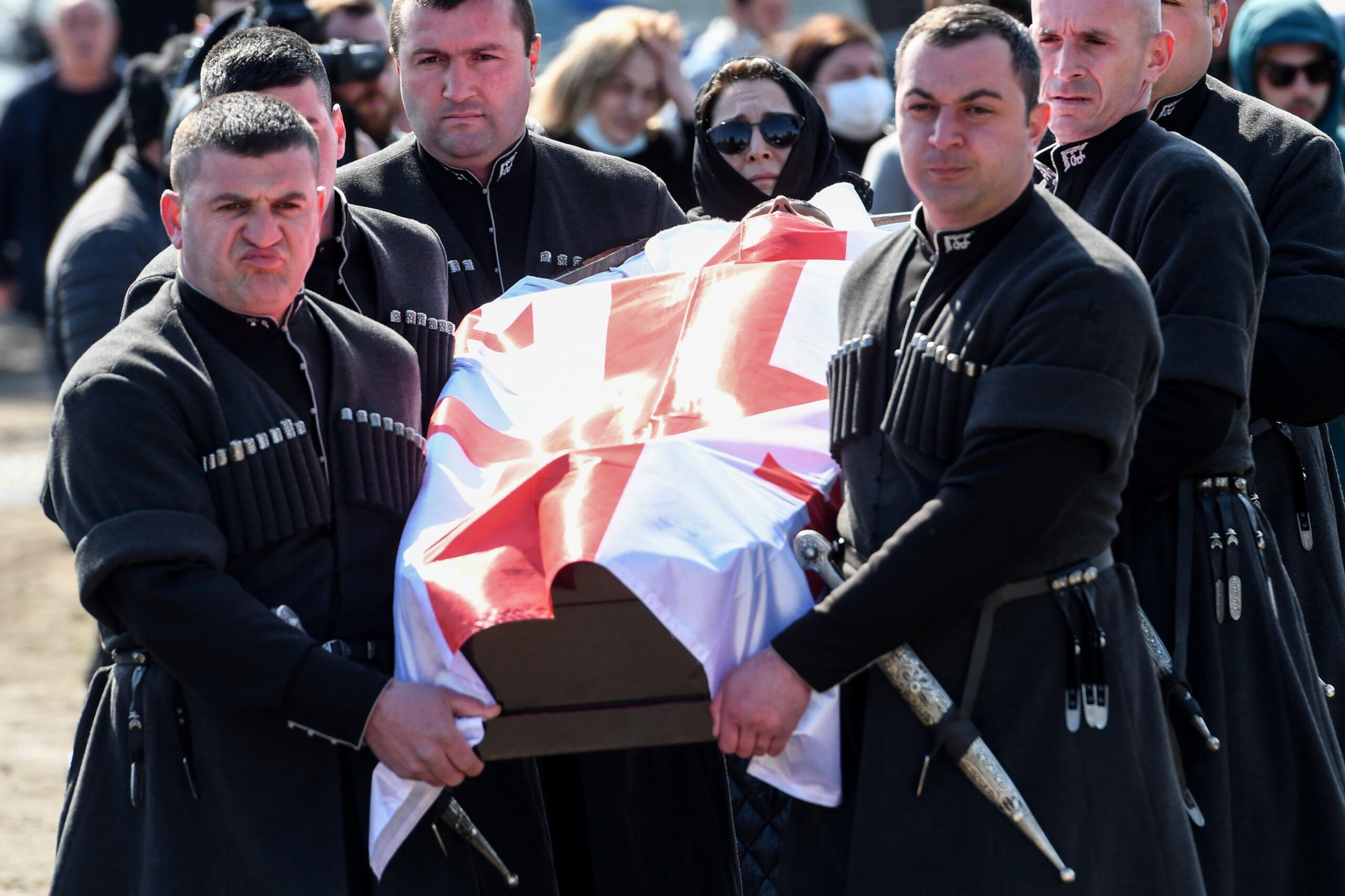 Mężczyźni w czarnych uroczystych strojach niosą trumnę na której jest gruzińska flaga