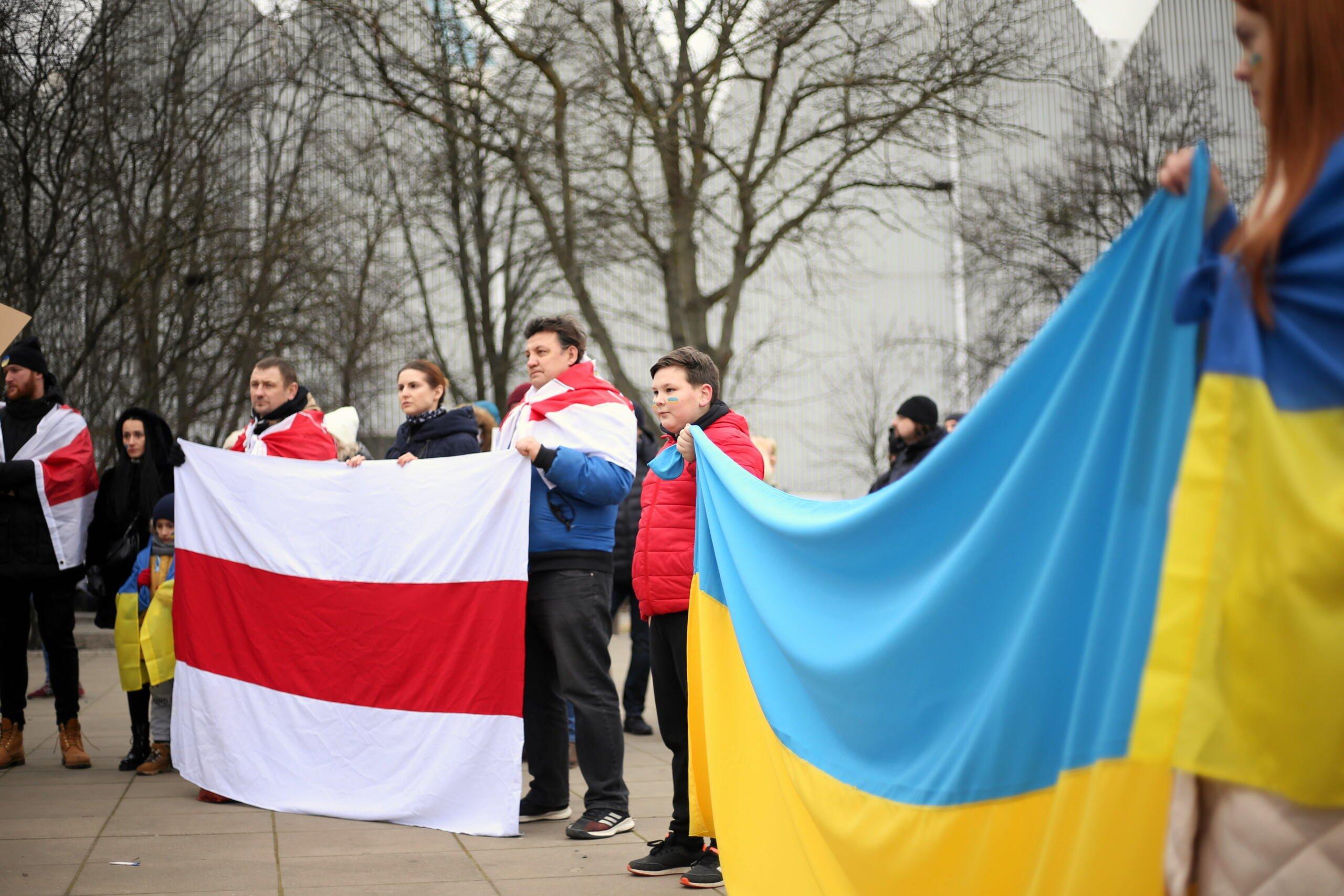 Ludzie na tlke drzew wparku stoją i trzymaja wielkie flagi Ukrainy i wolnej Białorusi