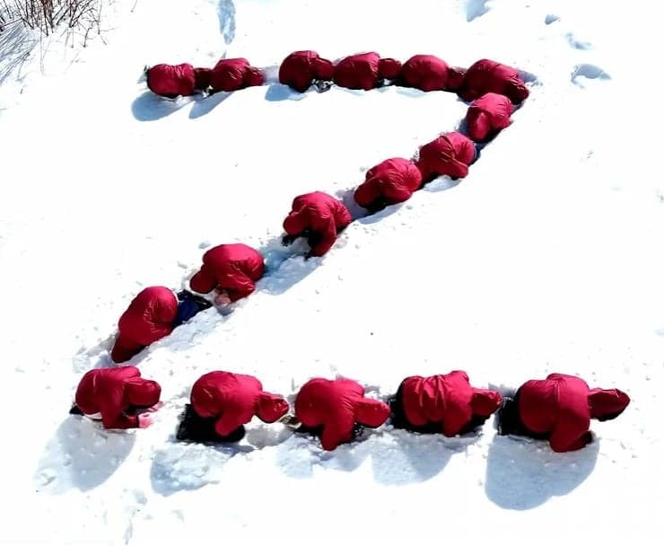 Dzieci ustawione w kształt litery Z w czerwonych kurtkach z kapturem klęczą z pochylonymi głowami w śniegu