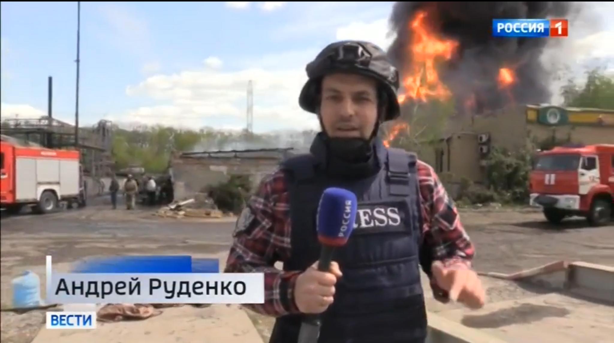 Dziennikarz w hełmie relacjonuje ogromny pożar widoczny w tle. Na mikrofonie napis ROSSIJA