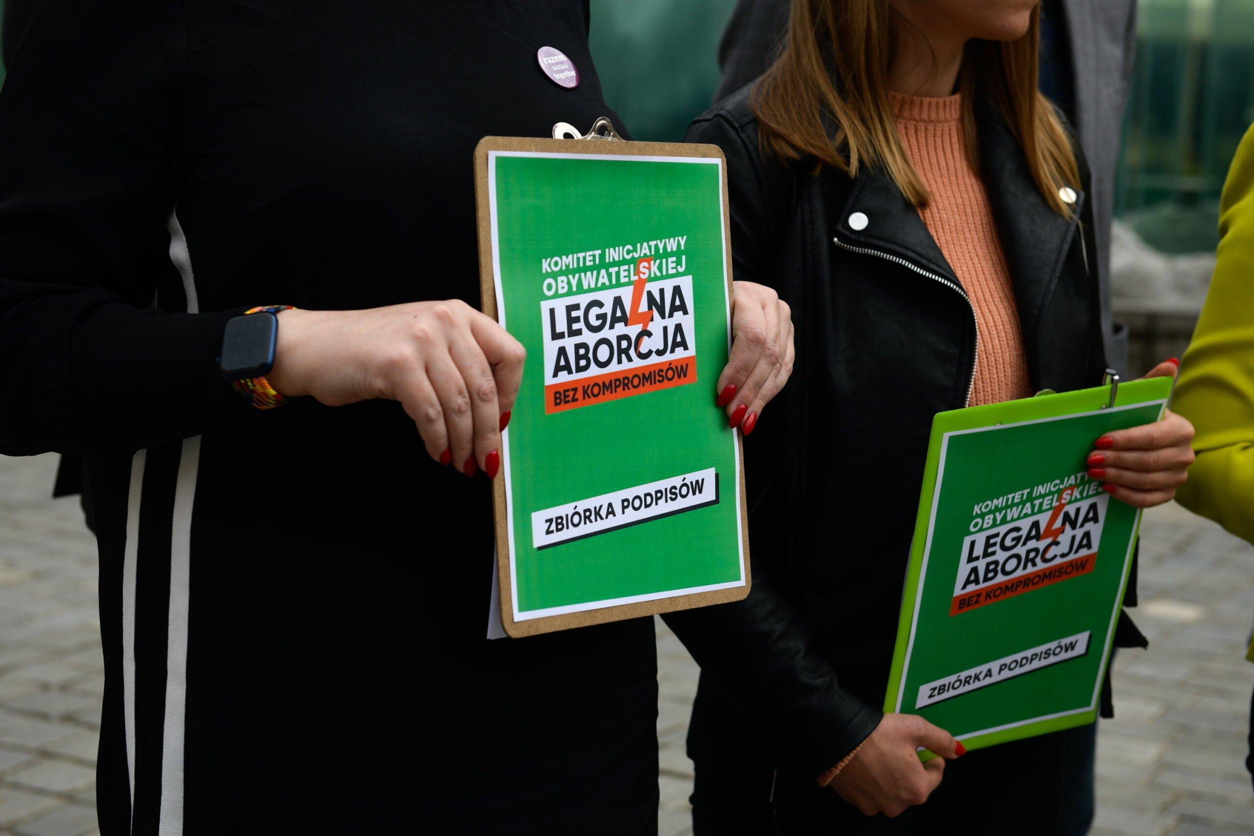 Dwie kobiety trzymają podkładki pod podpisy z hasłem "Legalna aborcja" na zielonym tle