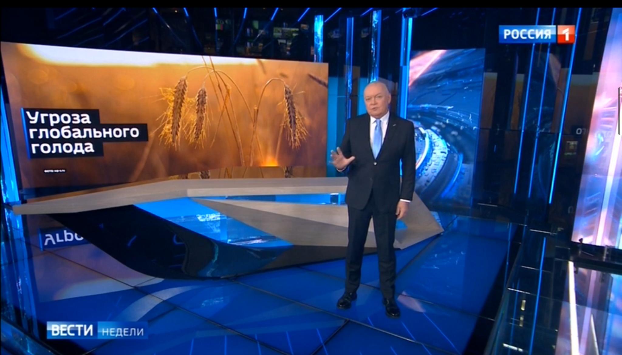 Studio telewizyjne, prezenter, w tle zdjęcie pola zboża i rosyjski napis "Zagrożenie klęską głodu na świecie"
