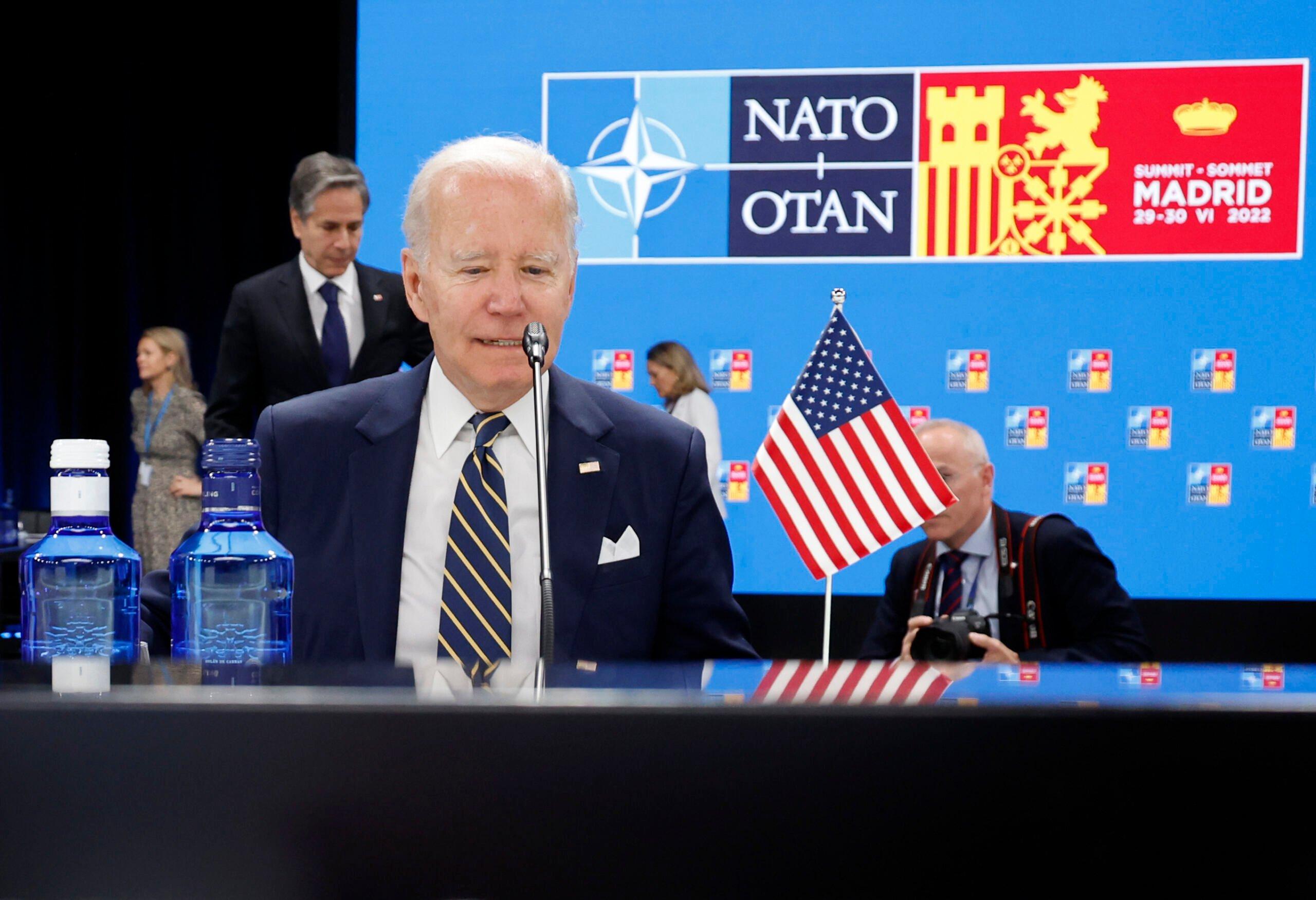 Jao Biden na tle logo NATO, siedzi przy stole z mikrofonem, obok flaga USA