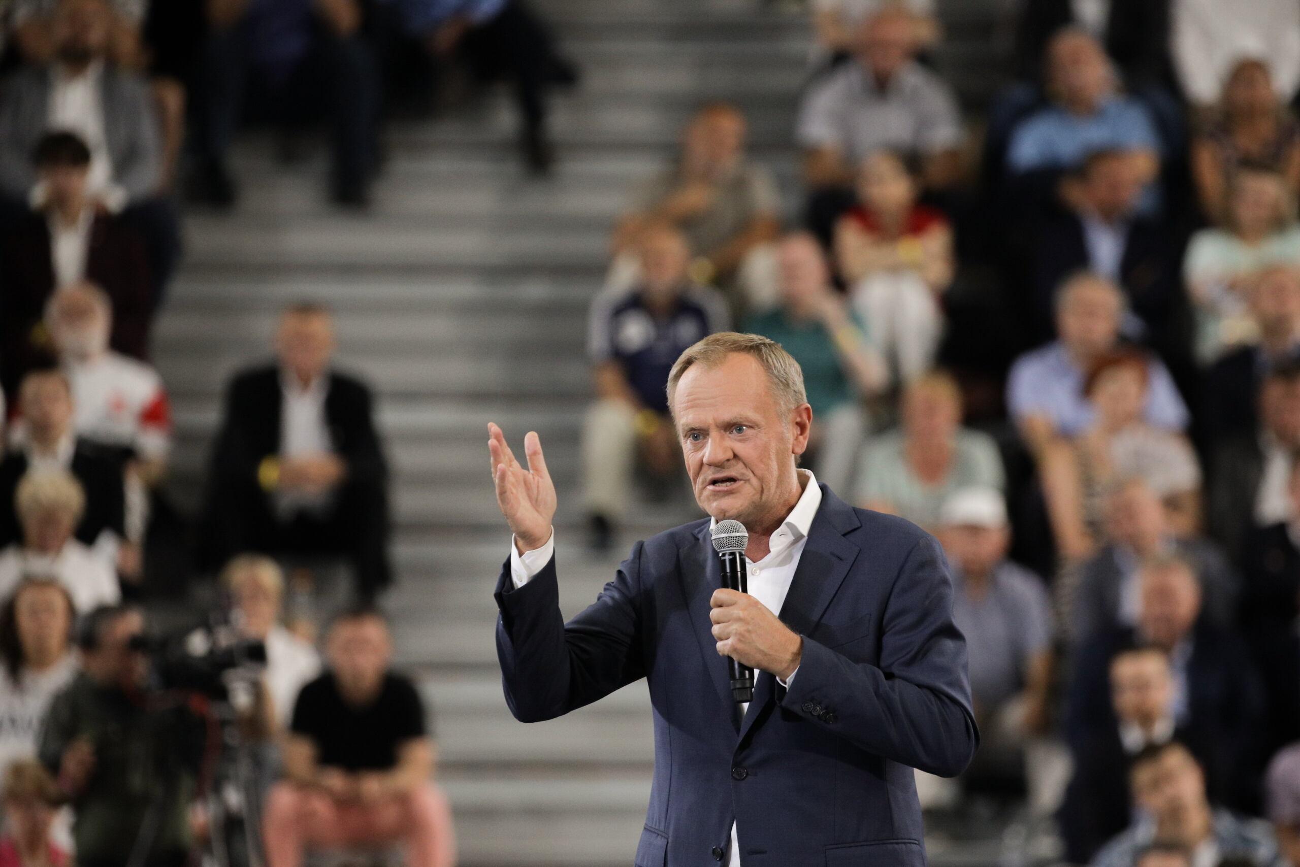 Donald Tusk z wyciągniętą ręką na tle trybuny wypełnionej ludźmi