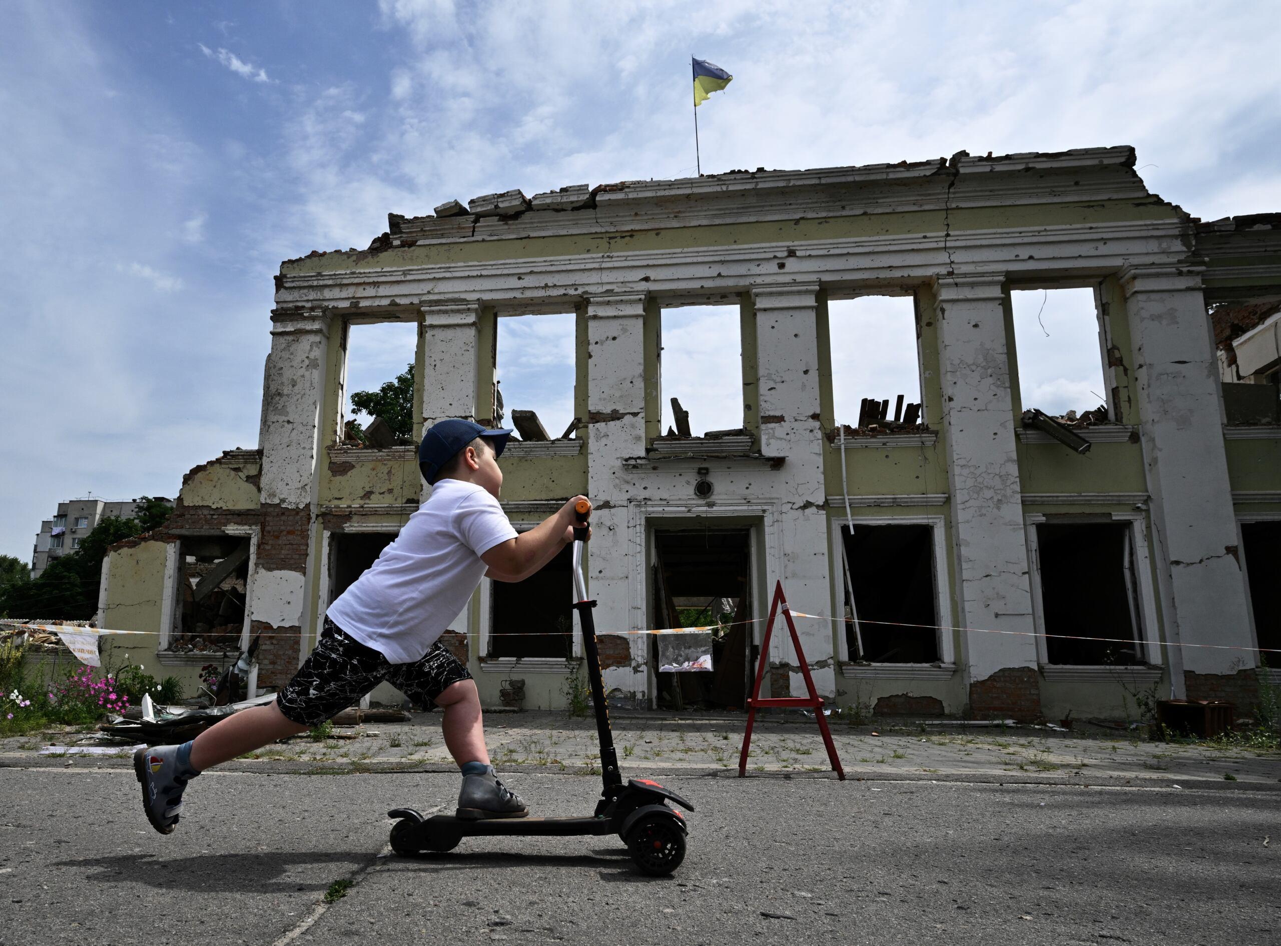 Chłopiec na hulajnodze, w tle zrujnowany budynek z ukraińską flagą