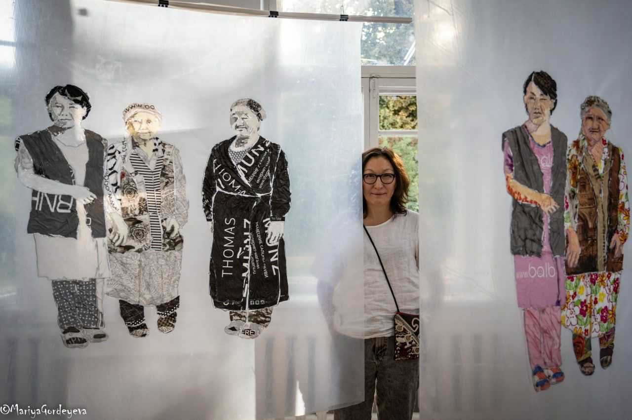 Kobieta w średnim wieku, przed nią płótna przedstawiające kobiety w tradycyjnych środkowoazjatyckich strojach